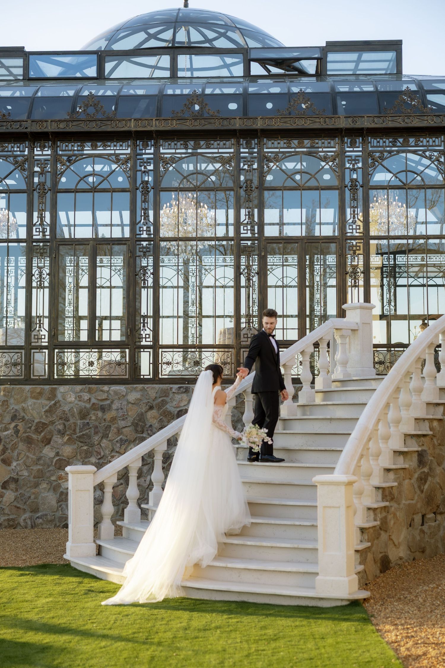 Bride and groom holding hands on elegant white stairs before a grand glass greenhouse venue.
