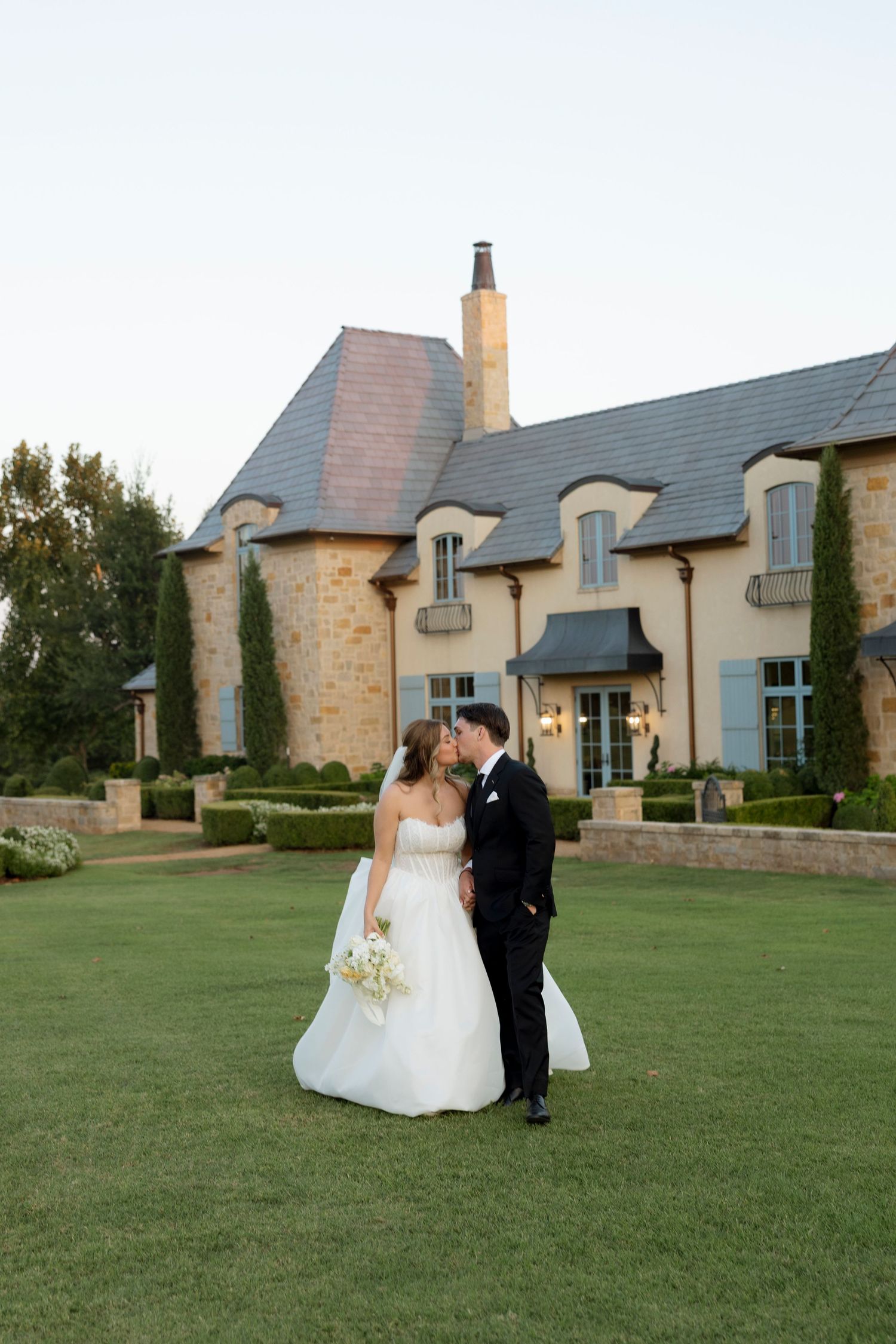 A wedding couple poses on a manicured lawn in front of a beautiful stone mansion with French architecture.