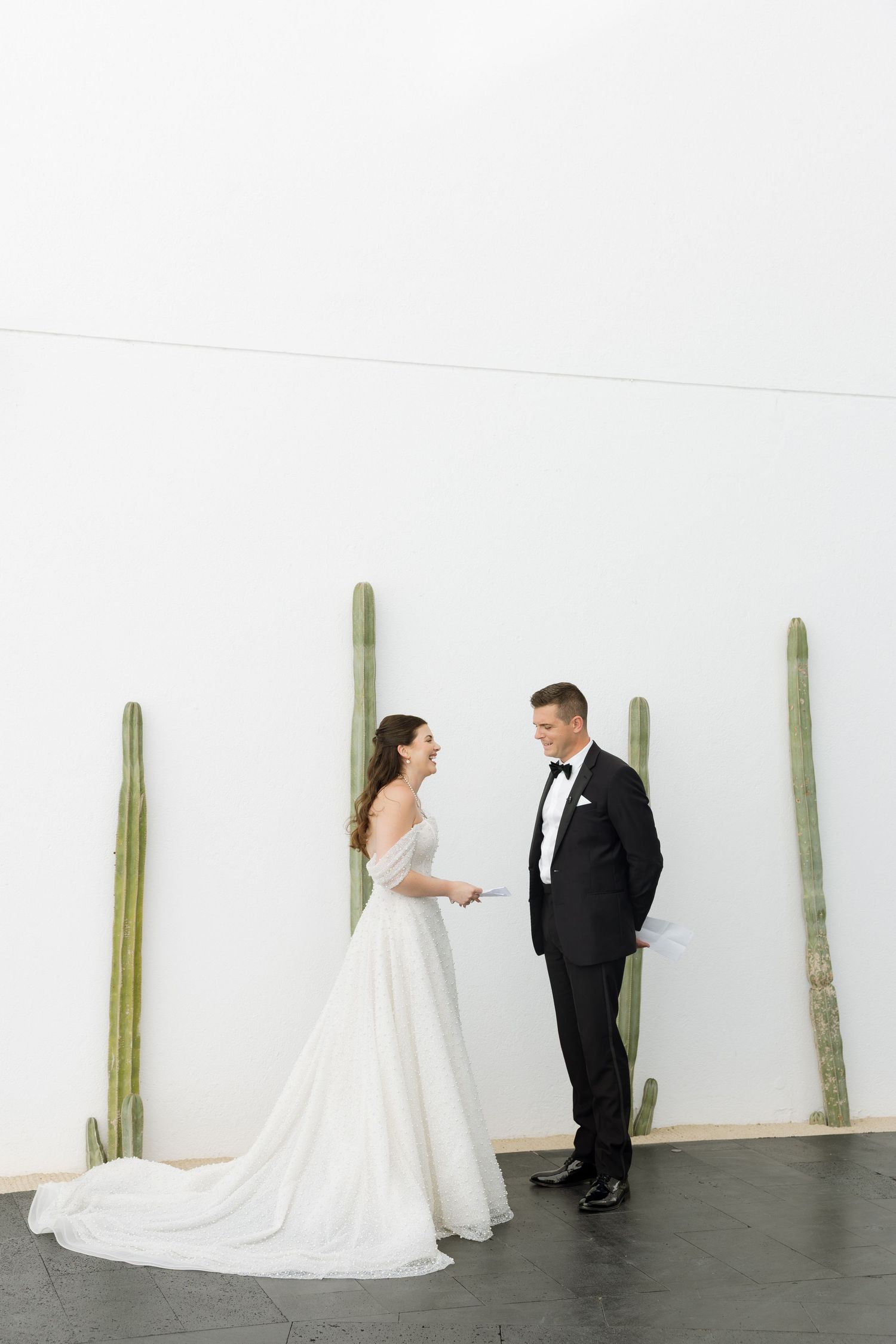 Bride and groom exchange vows in front of white wall with tall cacti at modern wedding venue.