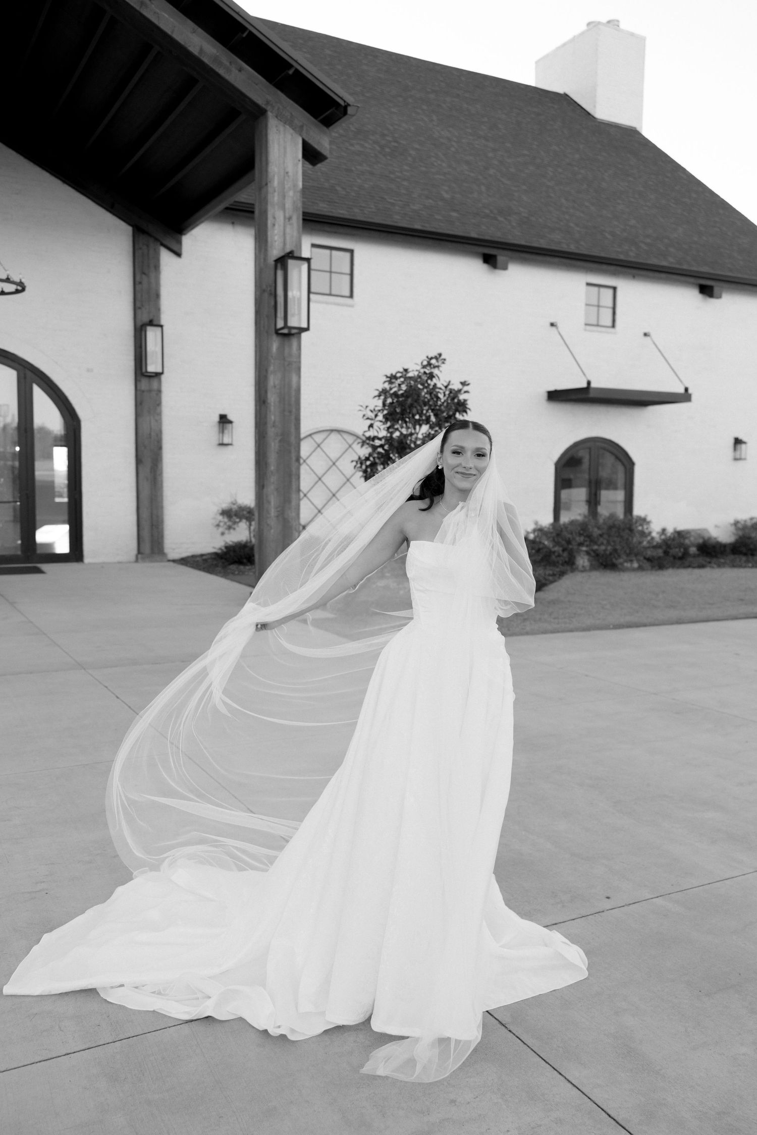A bride in a flowing white wedding gown with long veil poses outside a white stucco building in black and white.