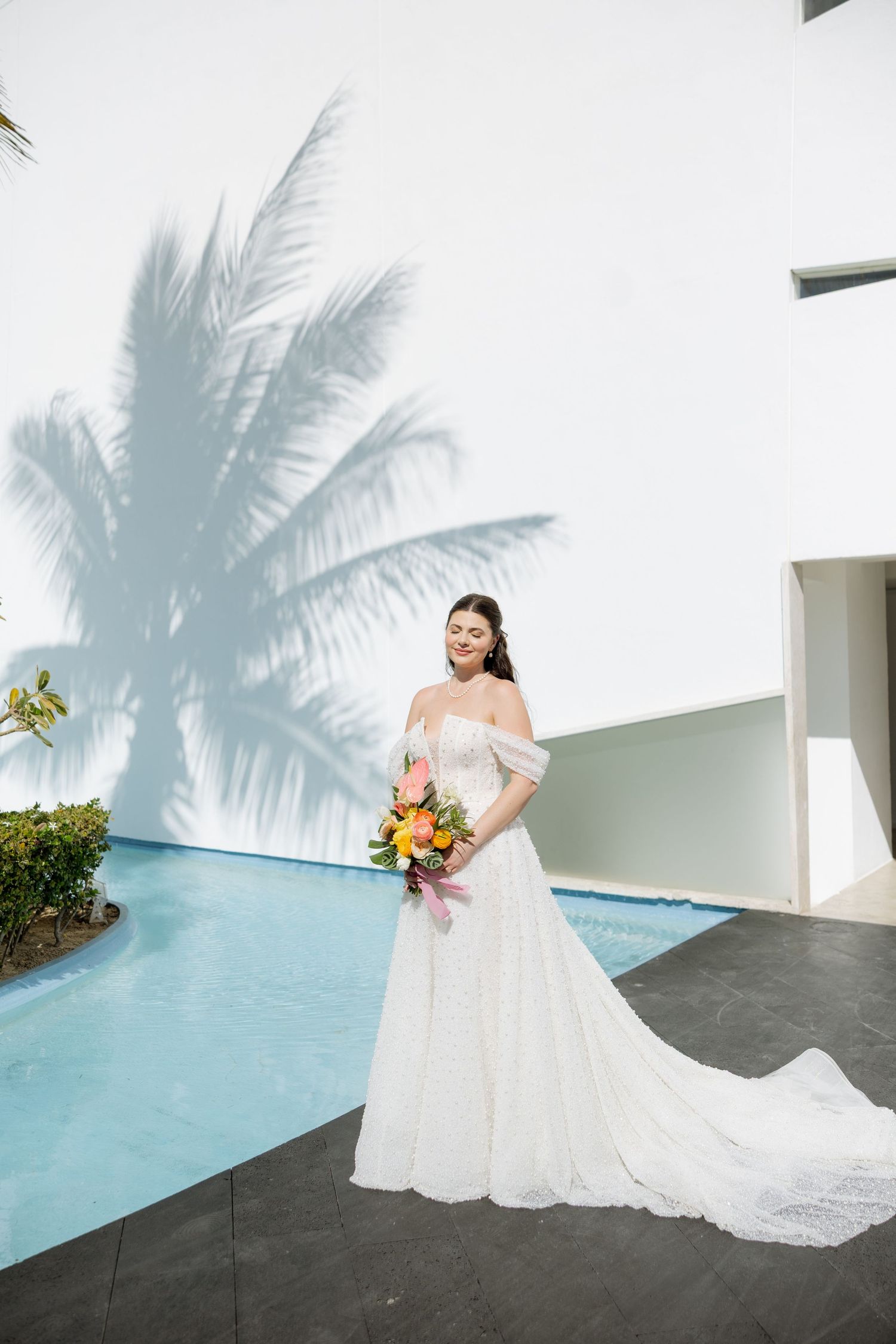 A smiling bride in a lace off-shoulder gown poses by a pool with a tropical bouquet.