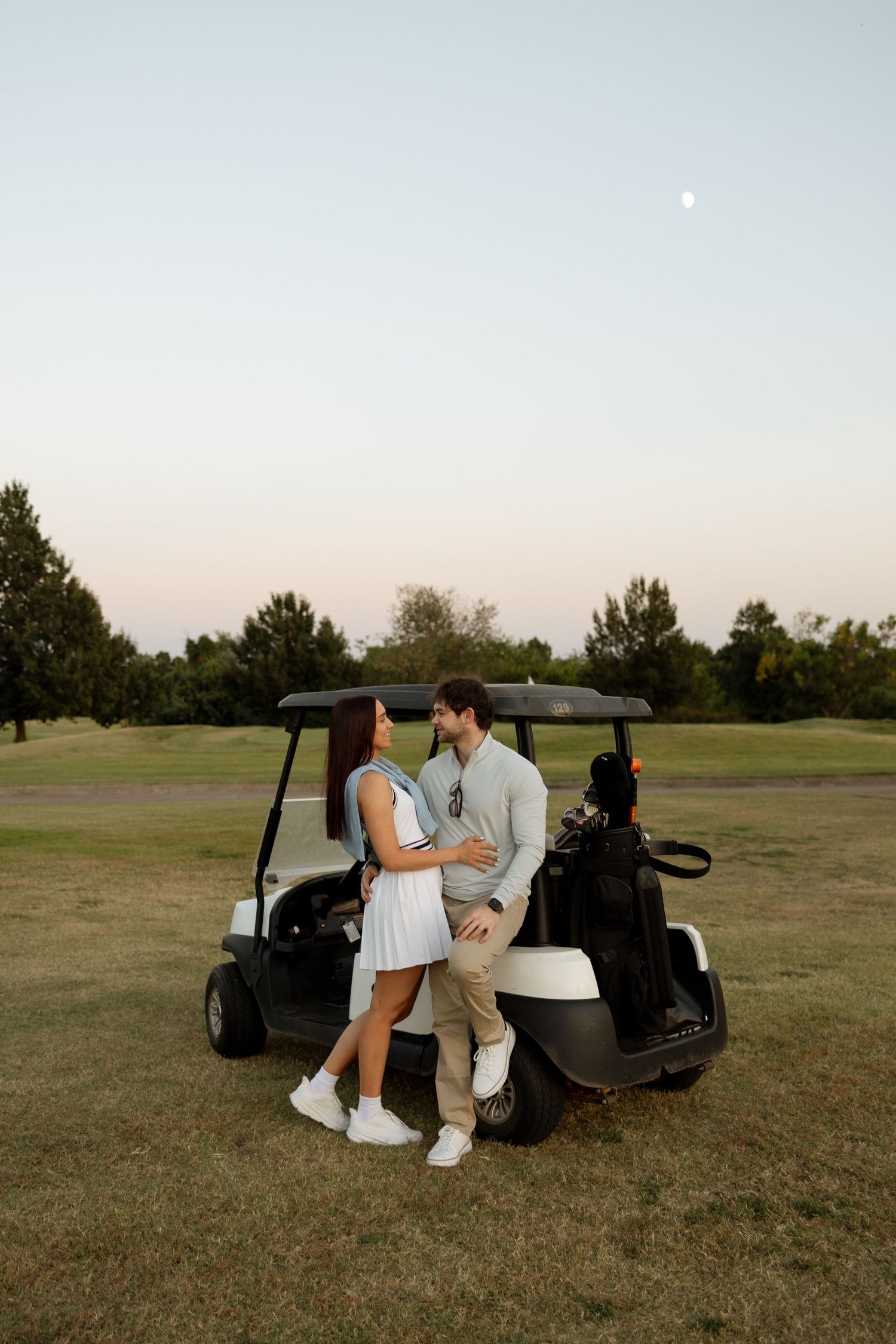 A romantic couple embraces next to their golf cart under a dusky evening sky.