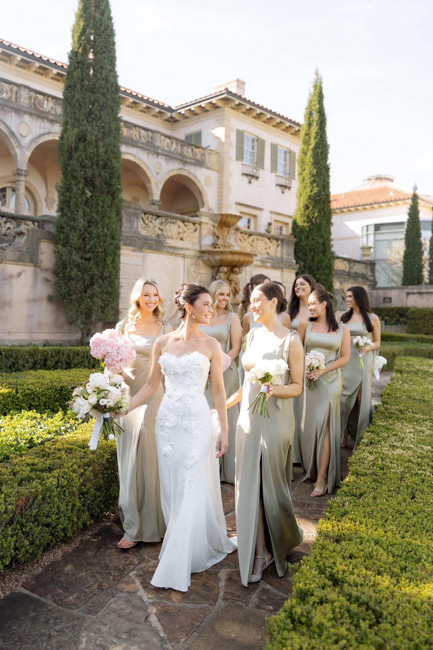 Bride in white lace gown walks with bridesmaids in sage green dresses through elegant garden venue.
