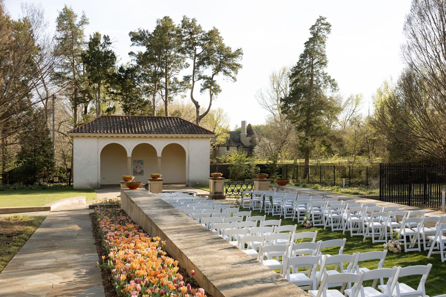 Outdoor wedding ceremony setup with white chairs, colorful flower beds, and an elegant arched pavilion surrounded by tall trees.