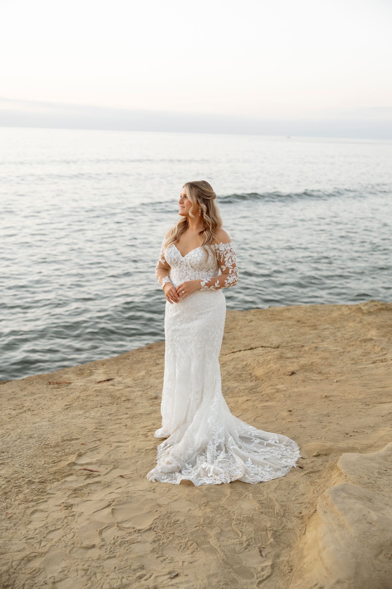 A bride in a lace wedding dress poses gracefully on a rocky beach at sunset.
