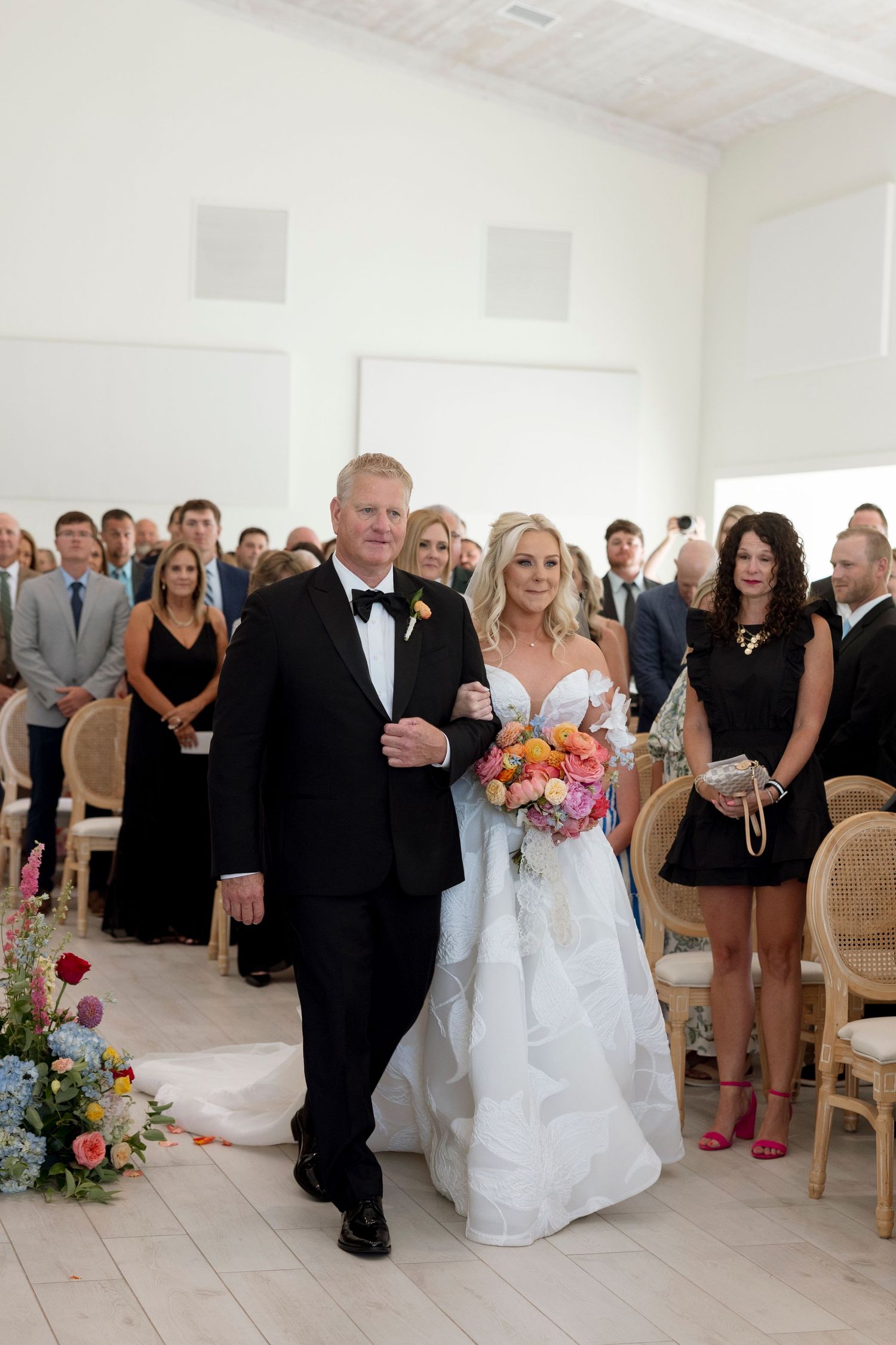 Wedding ceremony guests gather in an elegant white room with natural lighting.