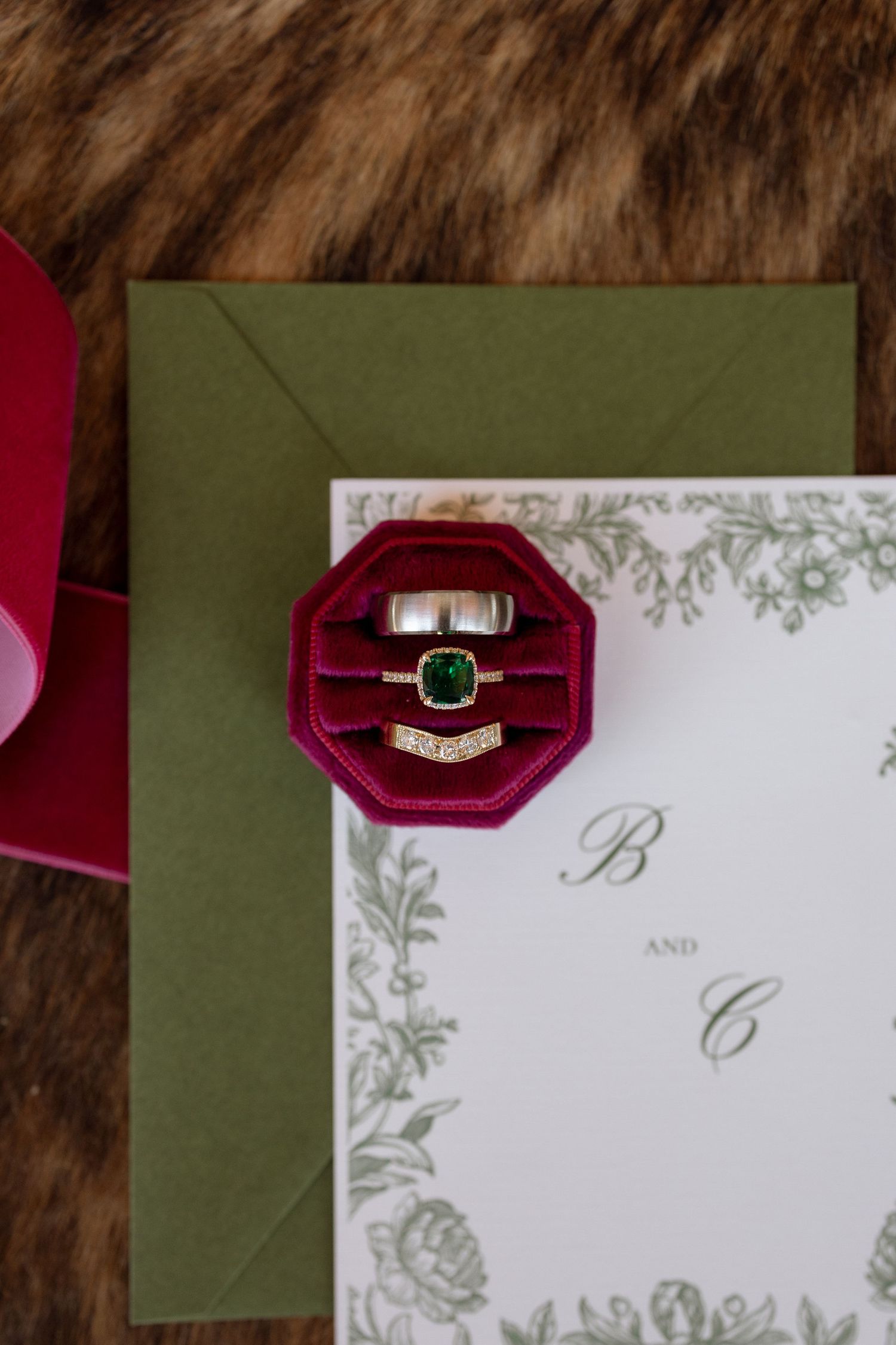 Wedding rings displayed in a burgundy velvet box on an elegant invitation with green details.