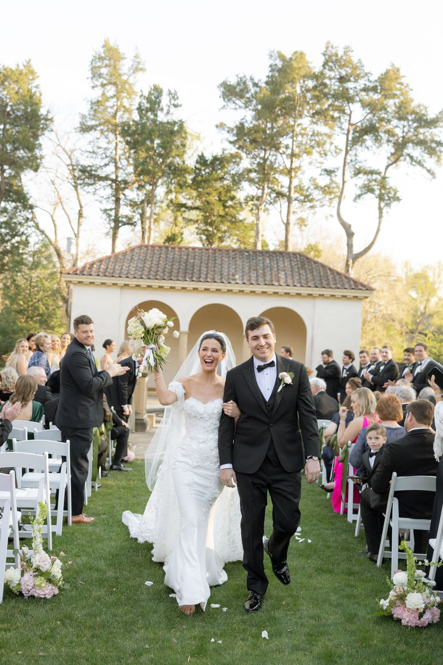 Beaming bride and groom walk back down aisle after ceremony at outdoor garden wedding with white chairs.