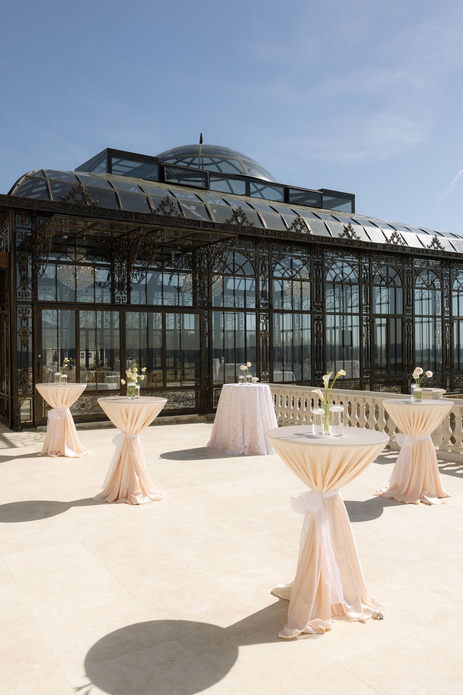 Elegant outdoor cocktail reception with draped bistro tables on a terrace in front of a Victorian glass greenhouse.