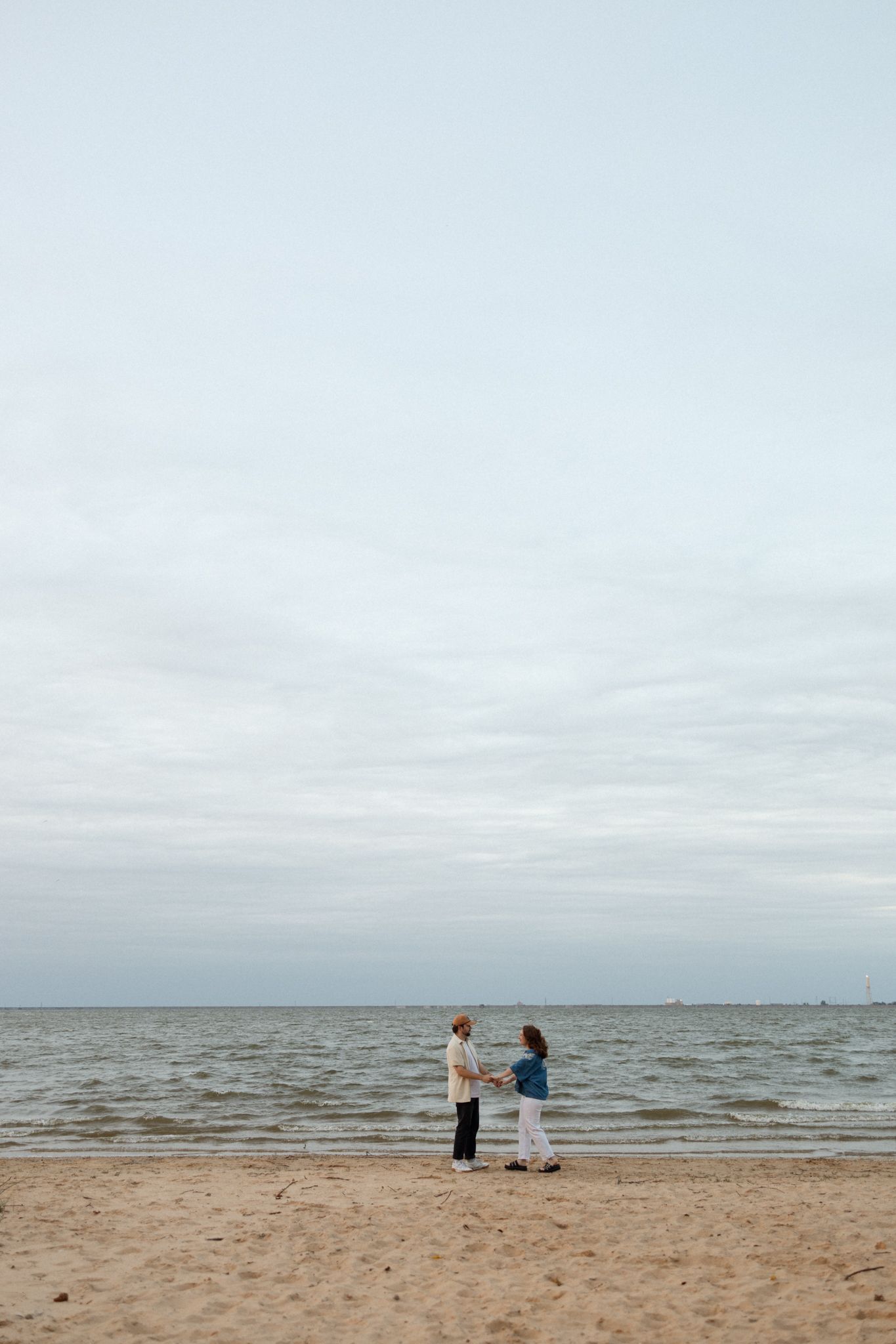 Two people stand together on an empty beach under an expansive cloudy sky.
