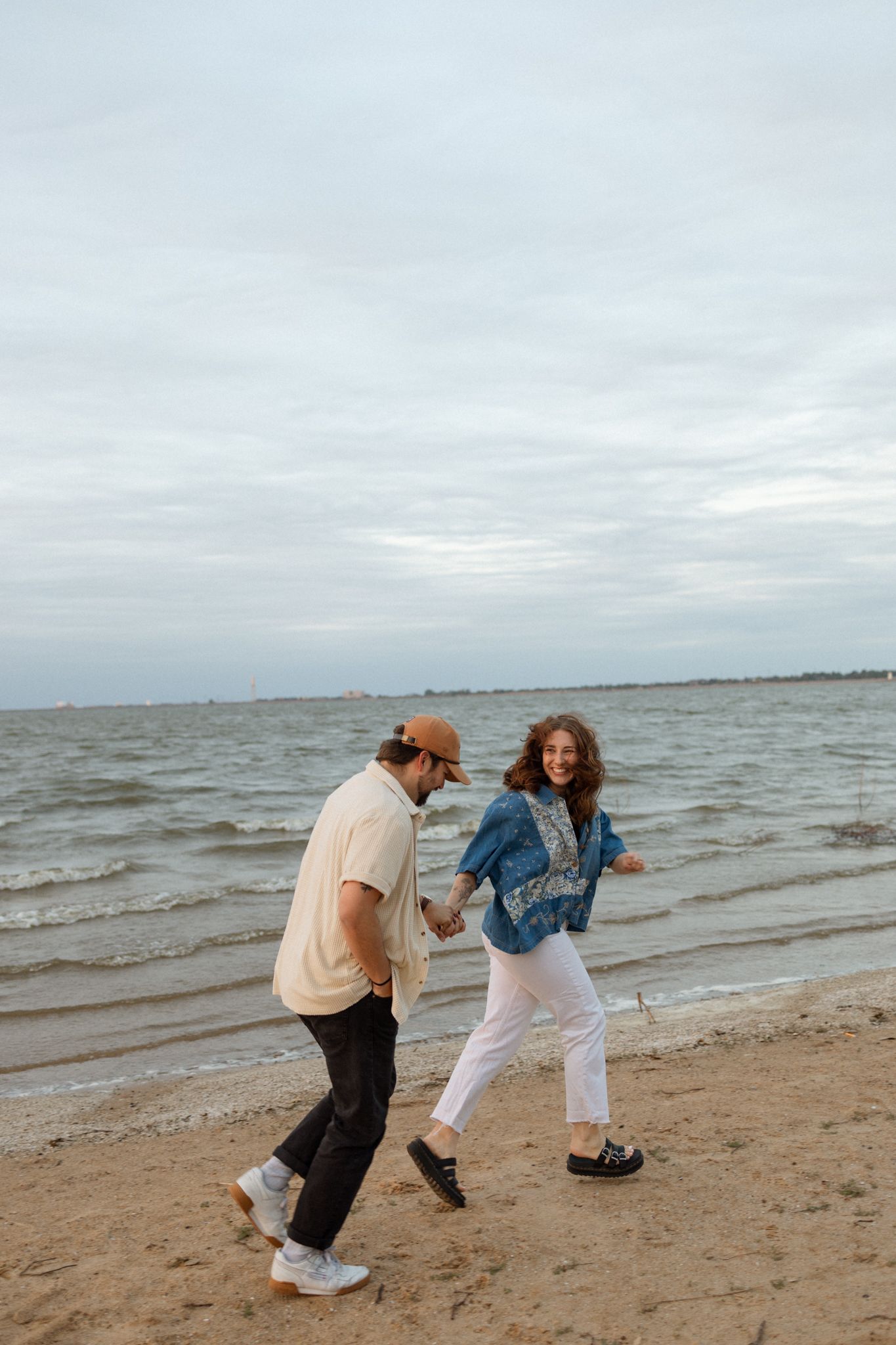 A playful couple runs along the beach holding hands under cloudy skies.