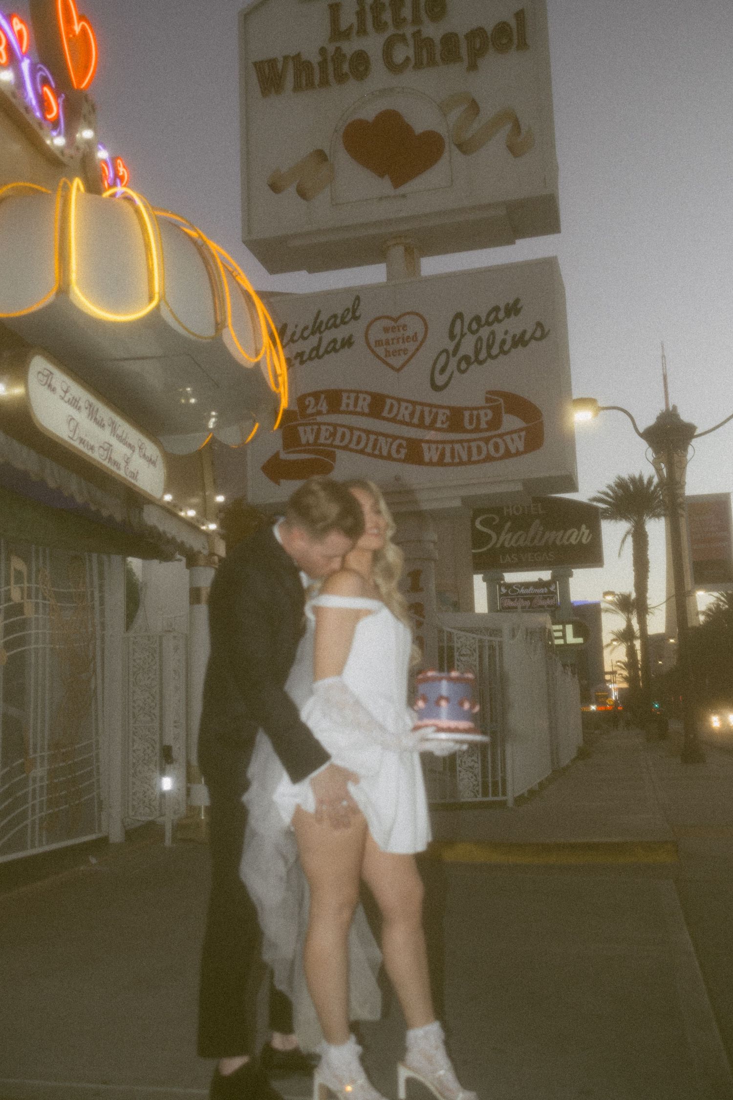 Couple shares intimate moment with wedding cake outside Las Vegas chapel at dusk.