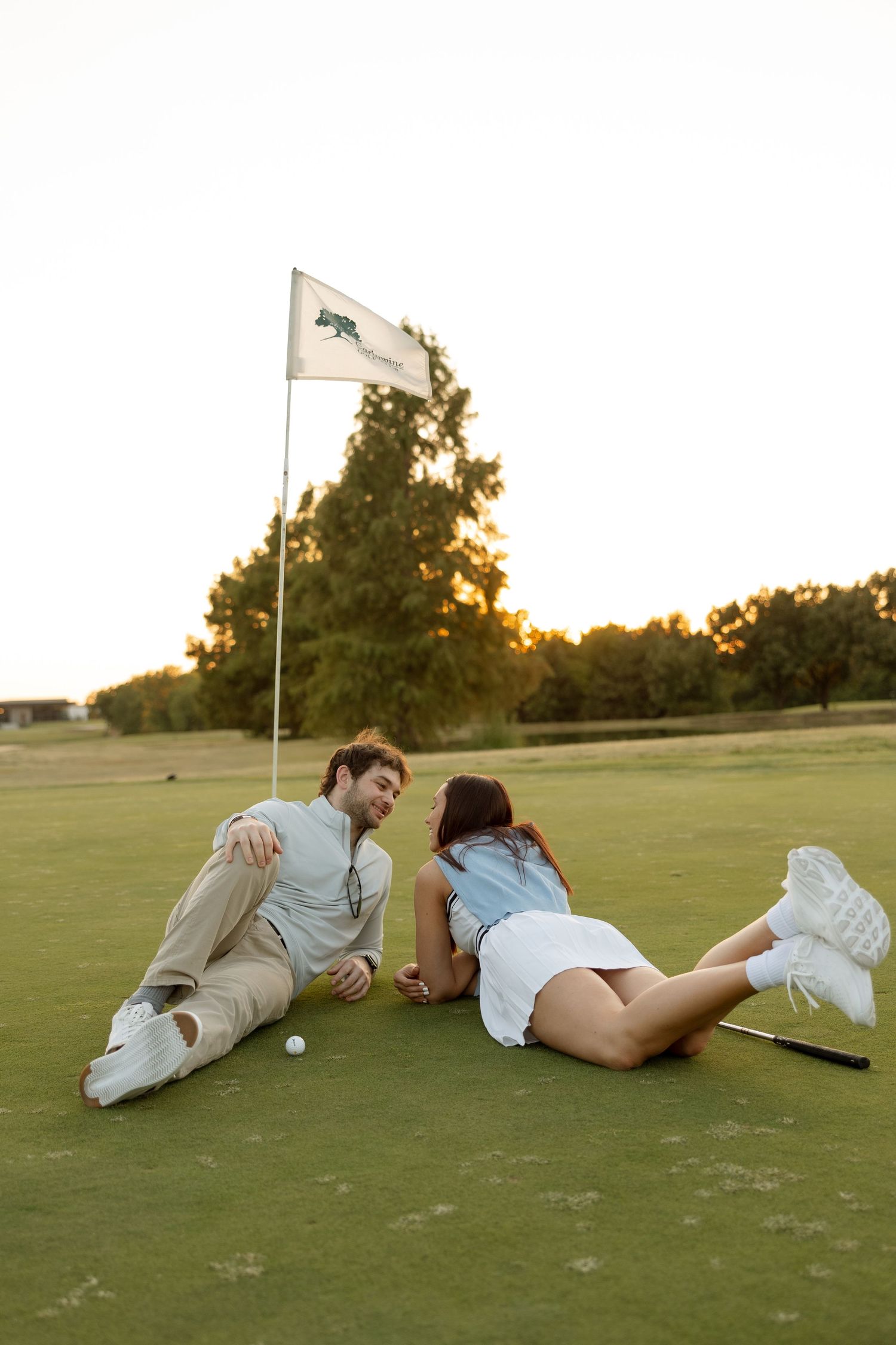 Couple relaxing on golf course grass near flag pole during sunset.