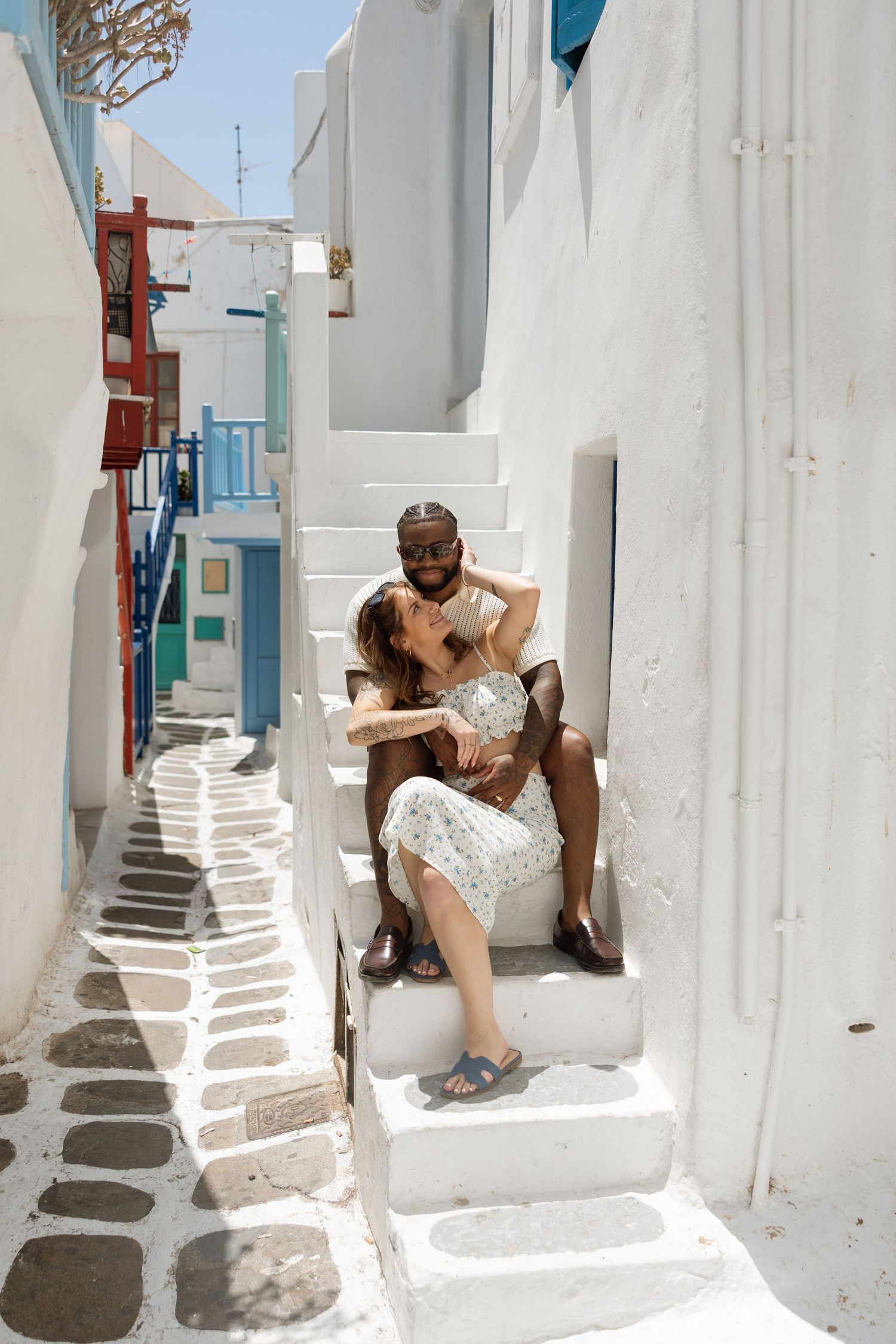 A couple sitting on white steps in a narrow traditional Greek alleyway with painted cobblestones.