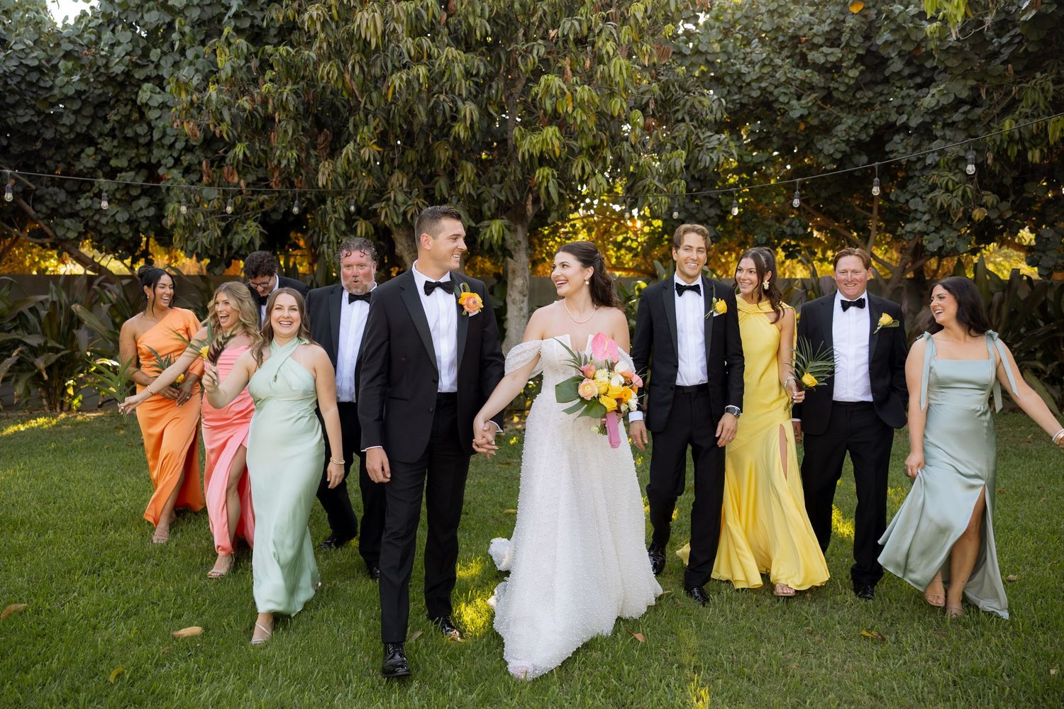 Wedding party walks outdoors in formal attire, bridesmaids in colorful gowns and groomsmen in black tuxedos with bride and groom.