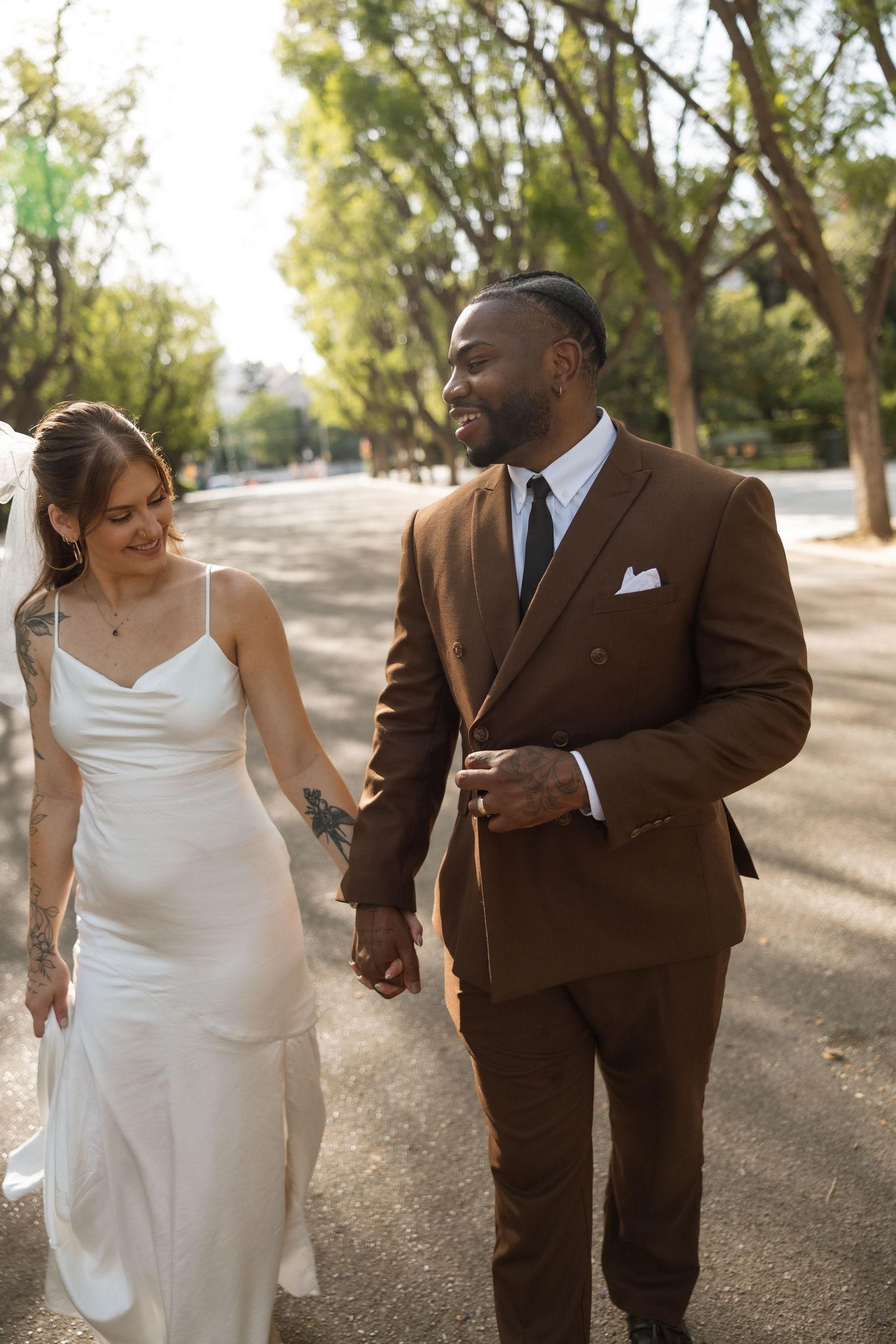 Newlyweds share intimate moment while walking along tree-lined street.