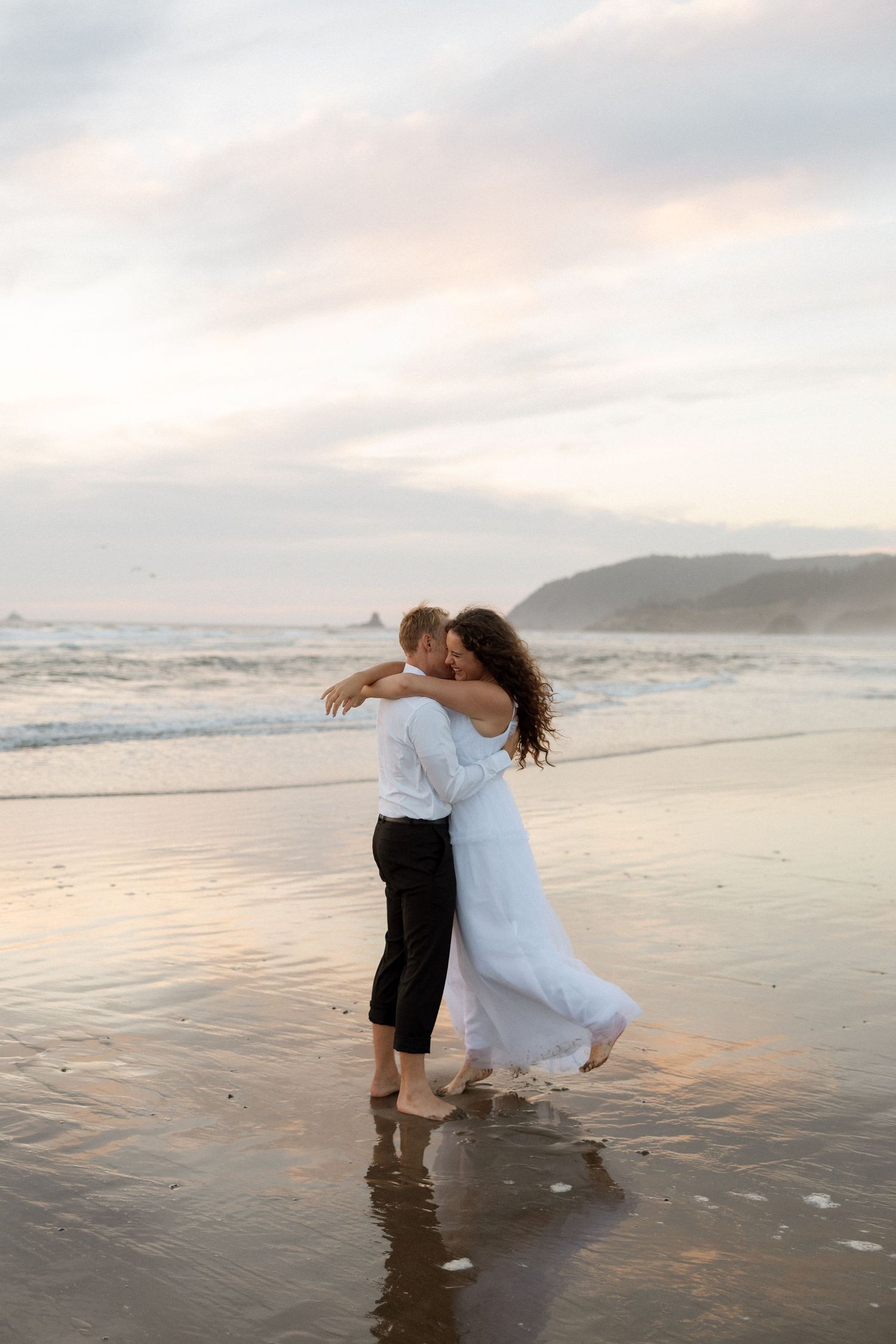 A couple dances on the wet sand with ocean waves and sunset clouds in the background.