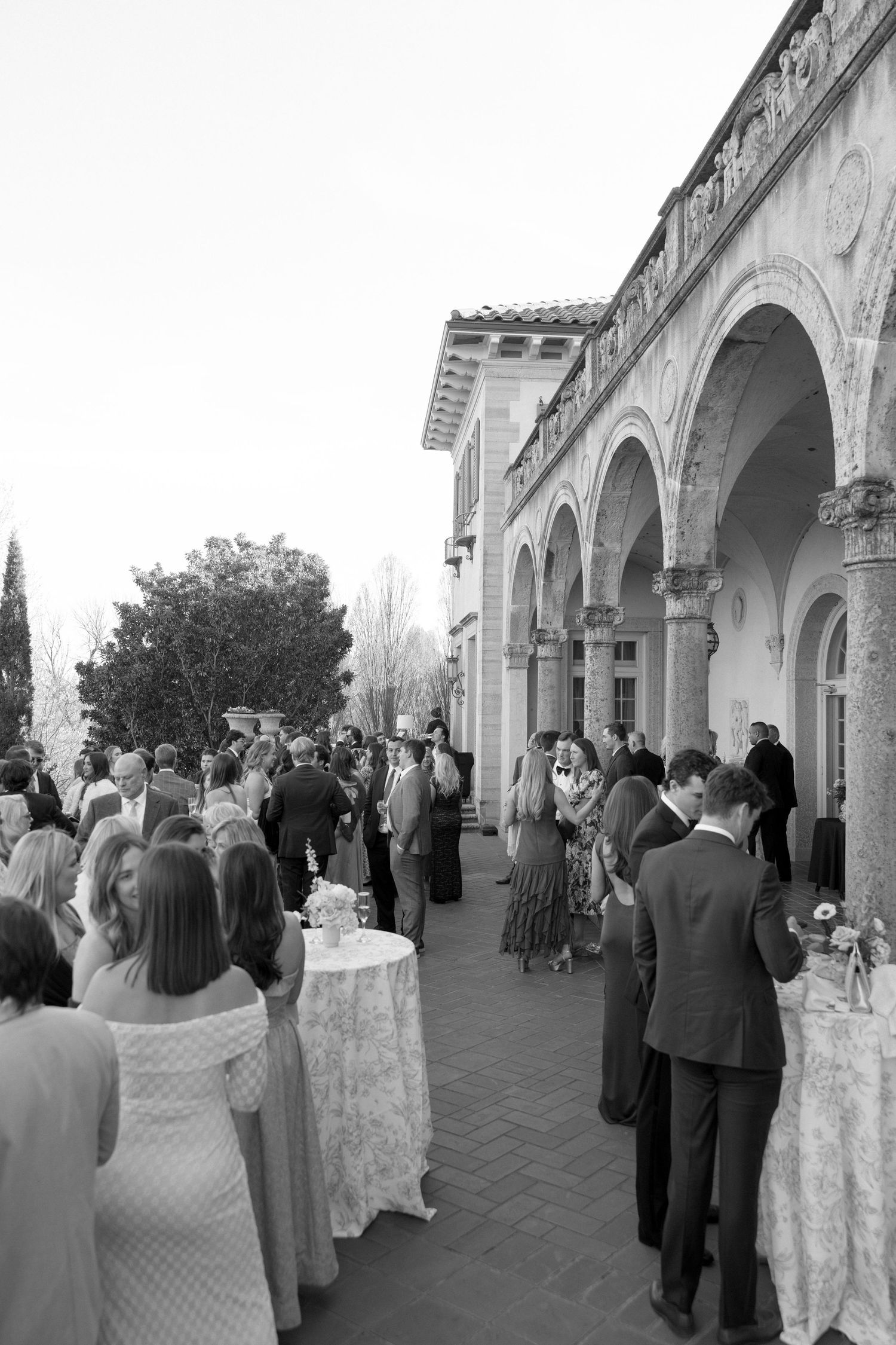 Elegant black and white photo of wedding guests mingling on a terrace outside a historic arched stone building.