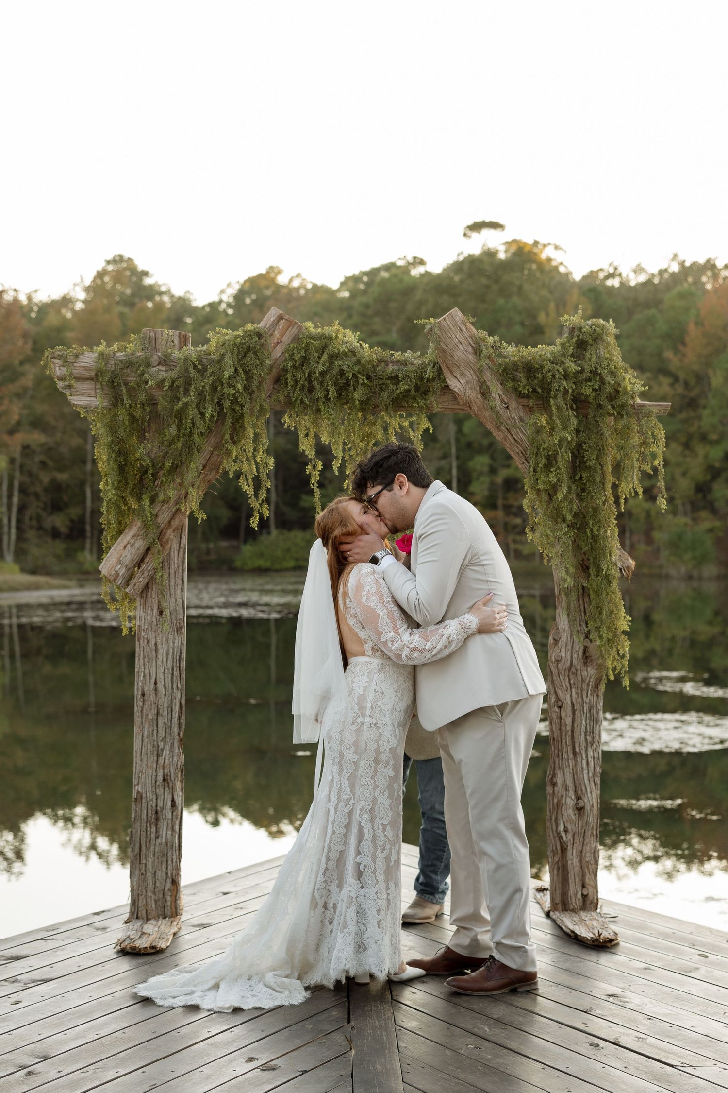 Newlyweds share their first kiss under a rustic arch decorated with moss by the lake.