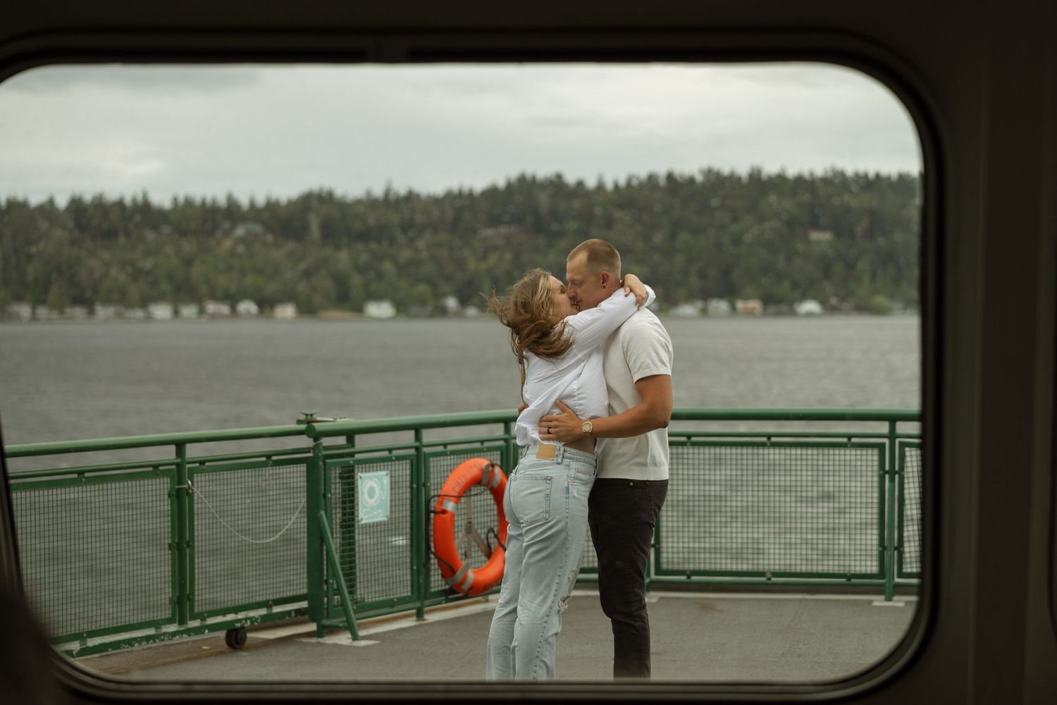 Couple embracing on ferry deck with Puget Sound waters and evergreen shore behind them.