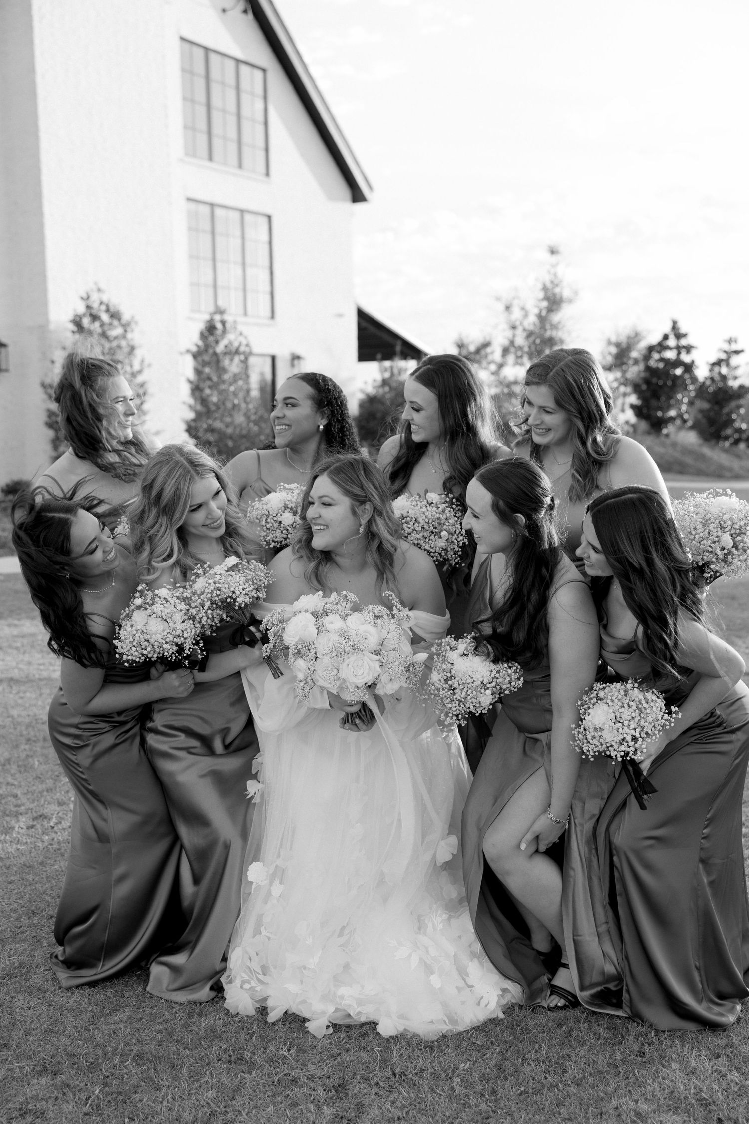 Bridesmaids in elegant mauve dresses gather around bride in white gown for joyful group photo outdoors.