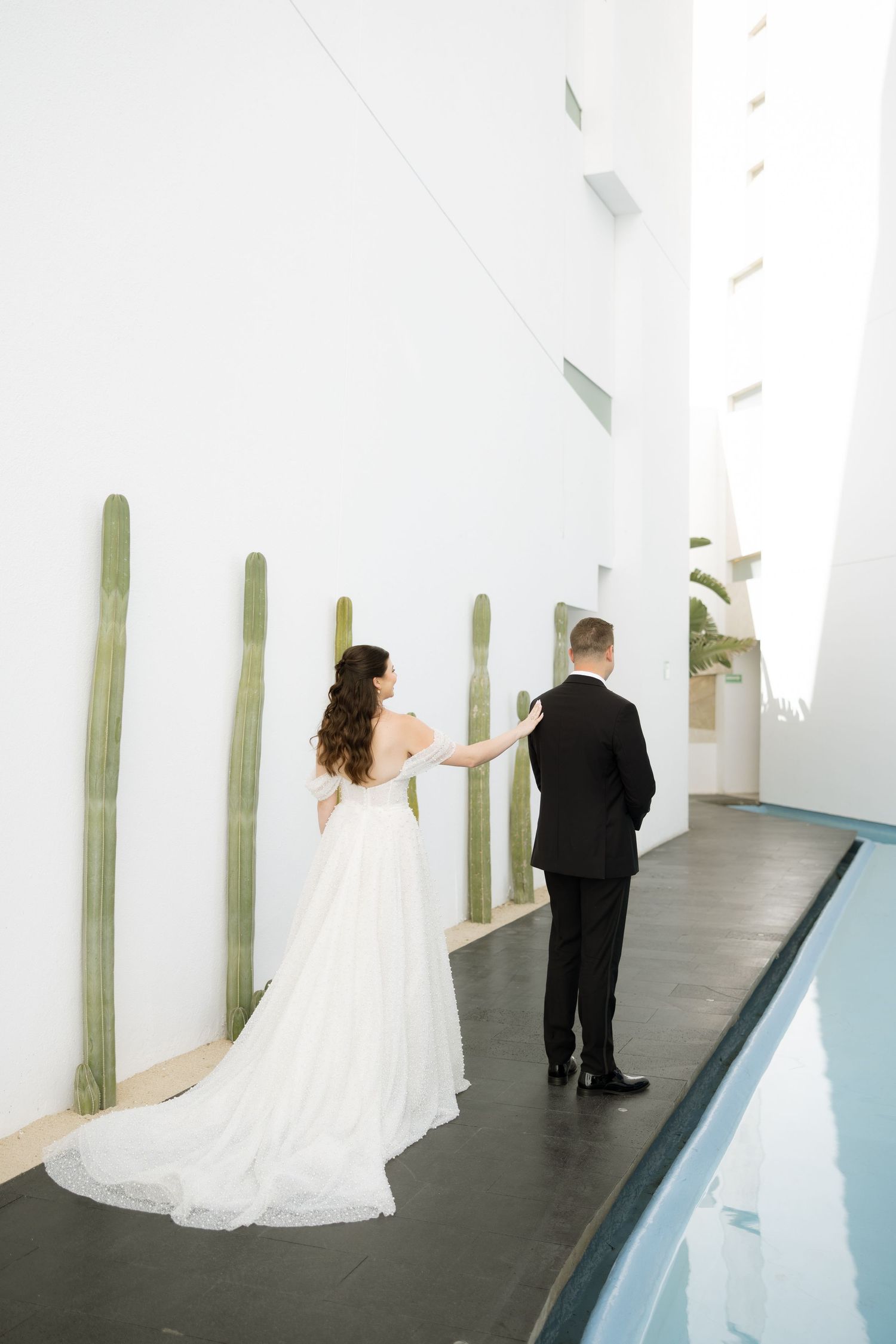 Bride in flowing white gown with long train and groom in black tuxedo walk along modern white wall with tall cacti.