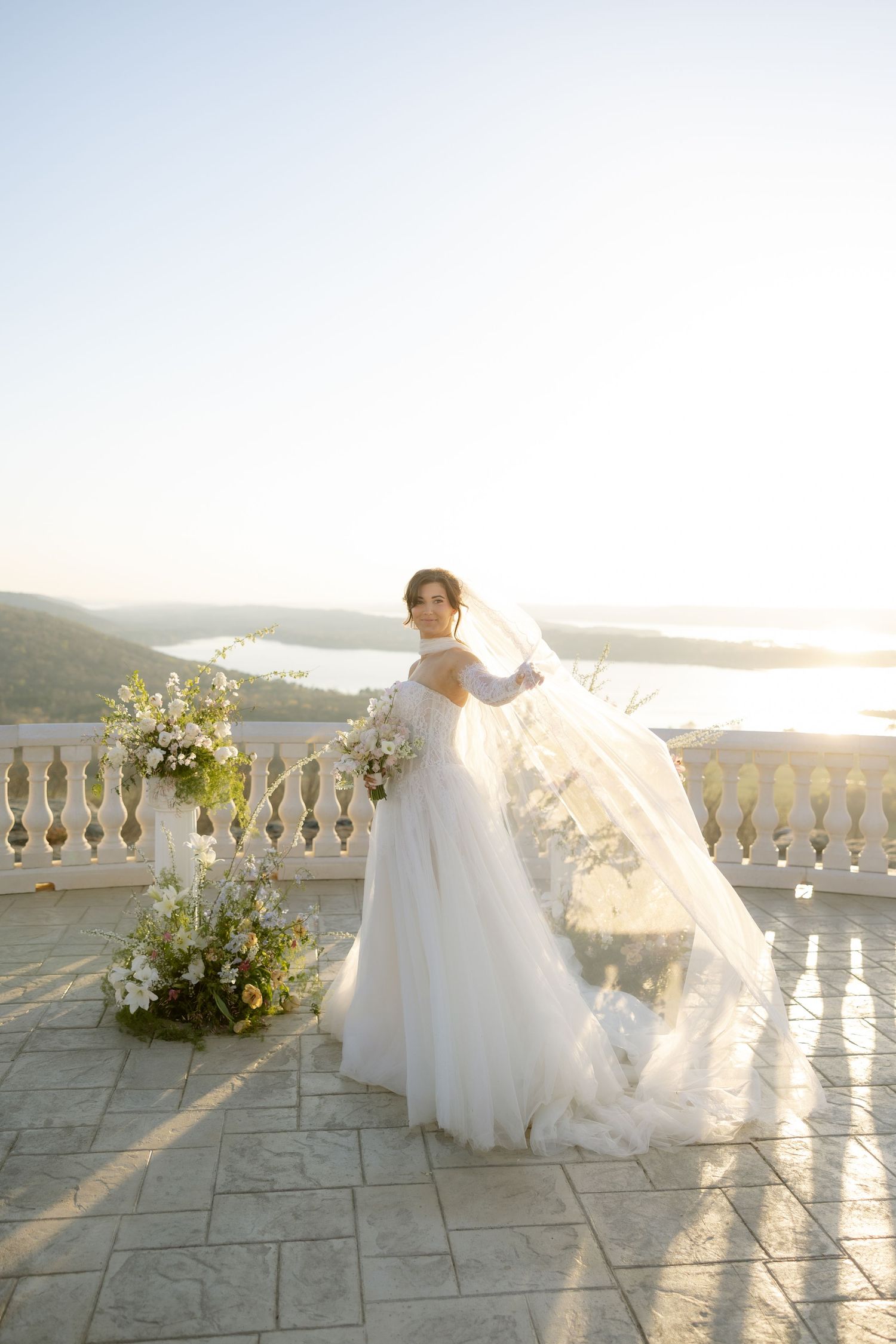 Bride in flowing white gown and veil stands on sunlit terrace with floral arrangements and scenic water views.