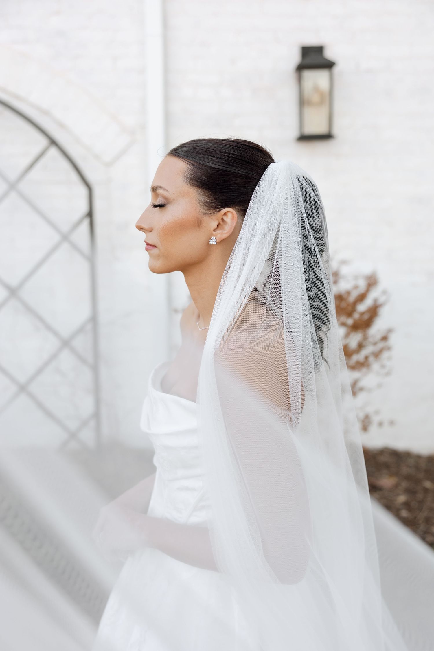Elegant bride in white wedding dress with flowing veil poses in profile against white wall with lantern detail.
