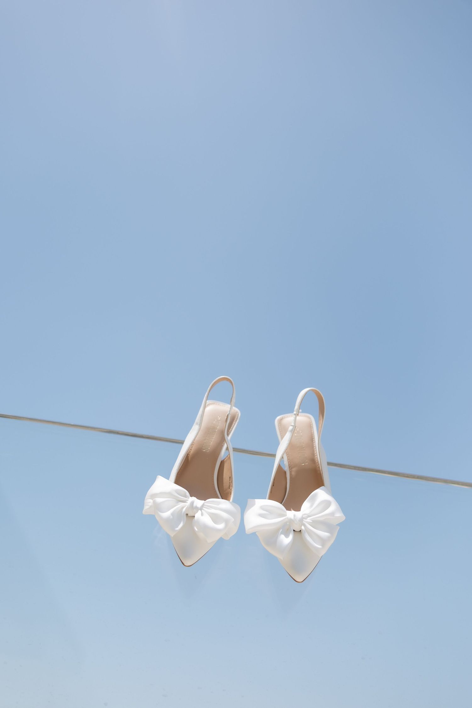 Elegant white wedding shoes with bows hanging on a clothesline against blue sky.