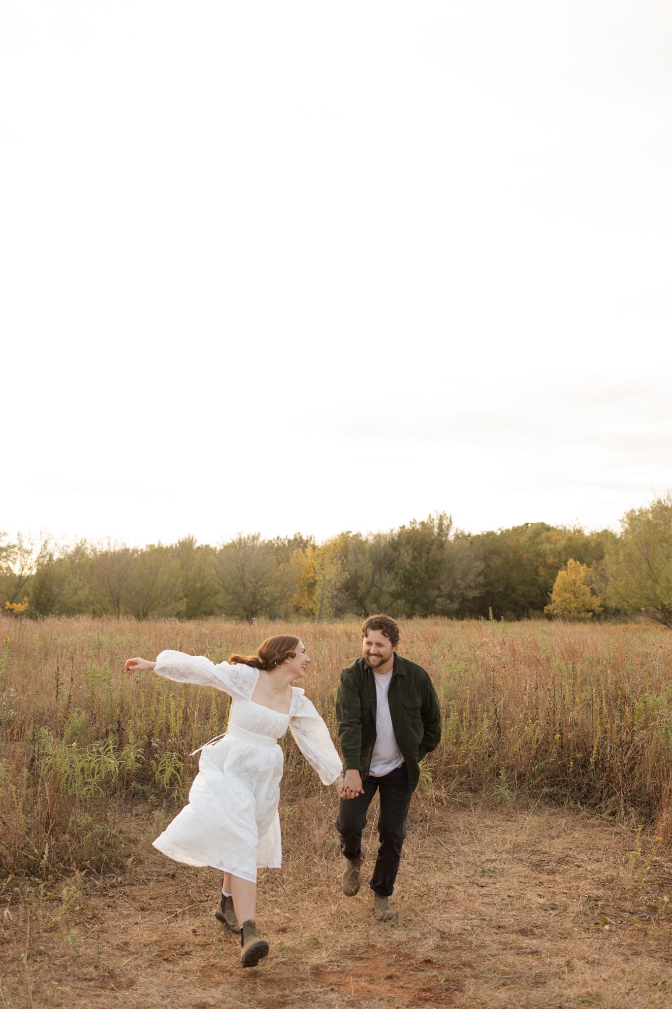 Couple in casual wedding attire runs playfully through an autumn field at sunset.