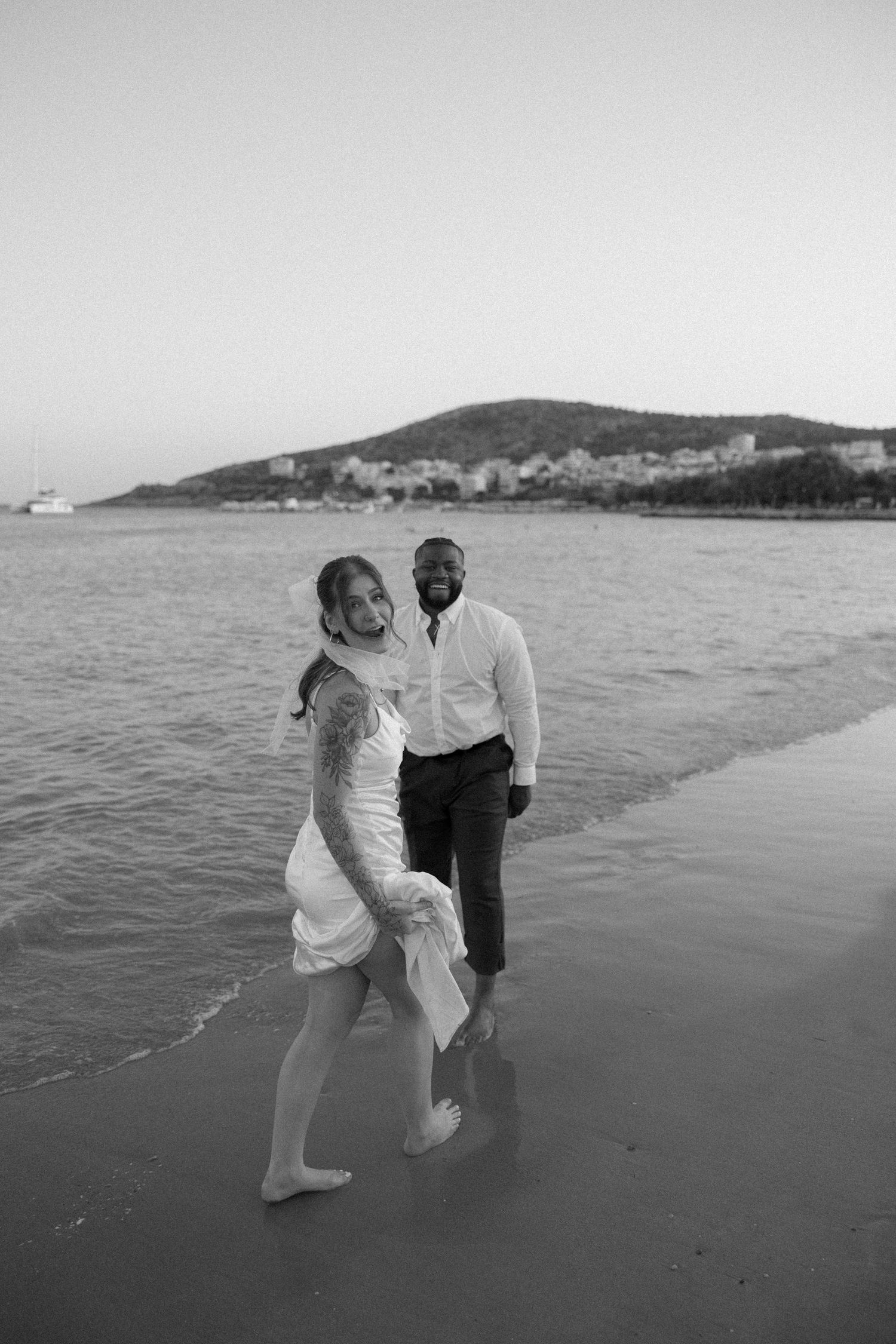 Black and white beach photo of couple enjoying playful moment by the water with coastal town backdrop.