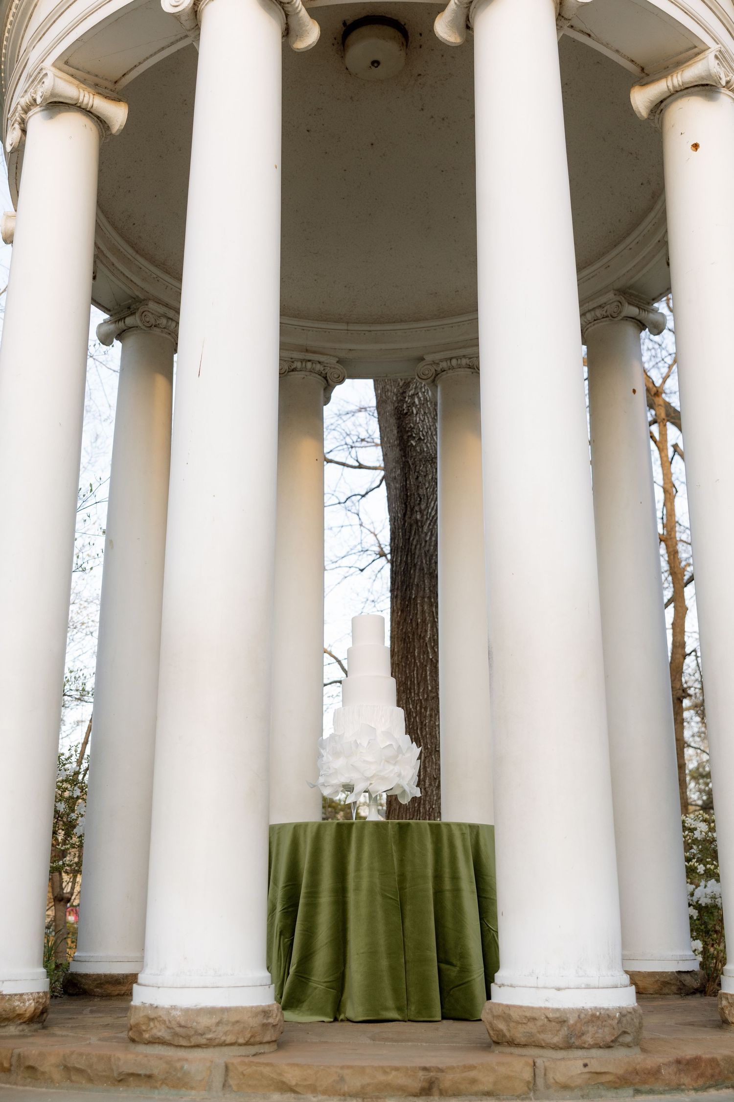 White columned rotunda with green draped table and white floral arrangement in an elegant outdoor wedding ceremony setting.