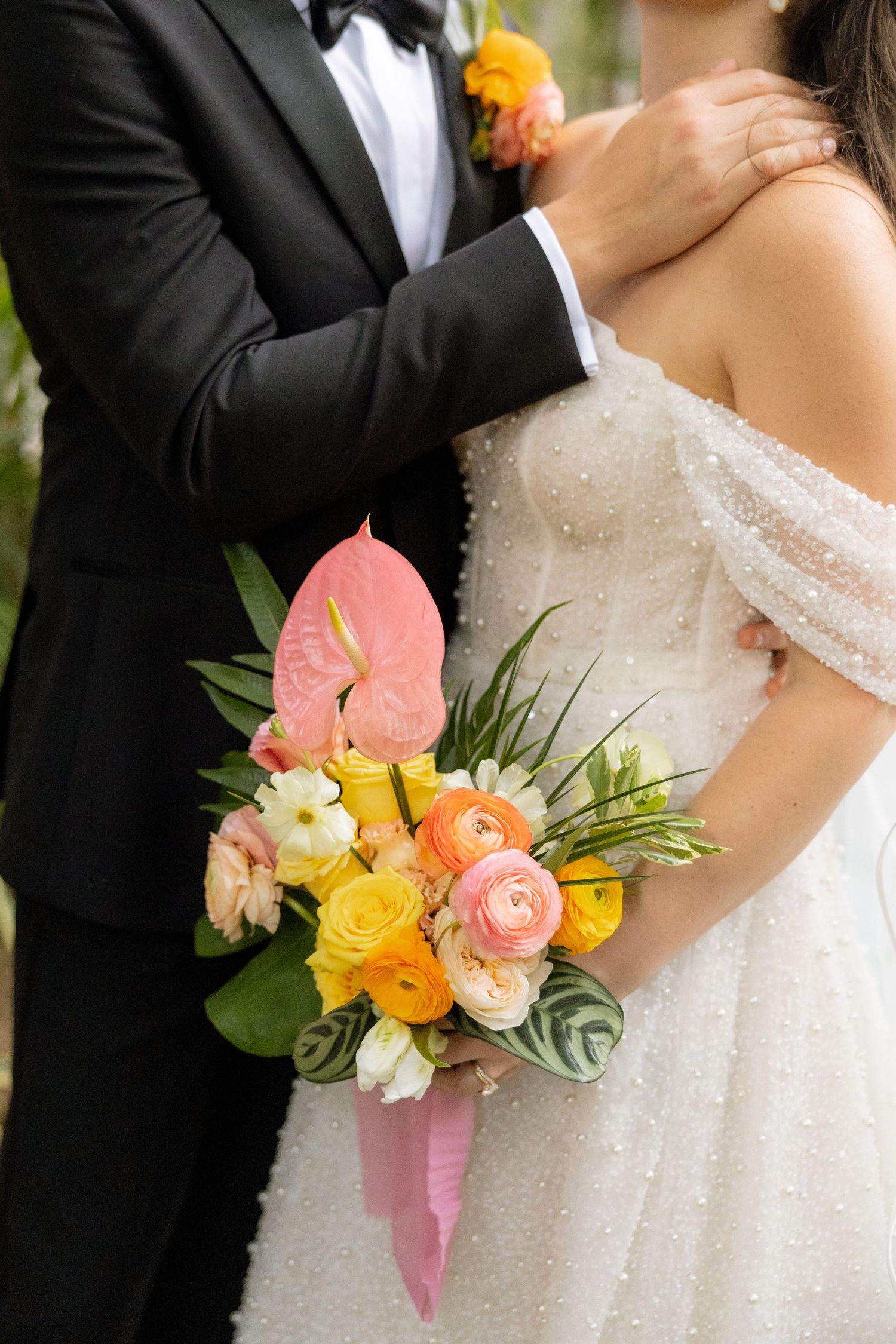 Bride holding tropical bouquet with pink anthurium and yellow ranunculus beside groom in tuxedo.