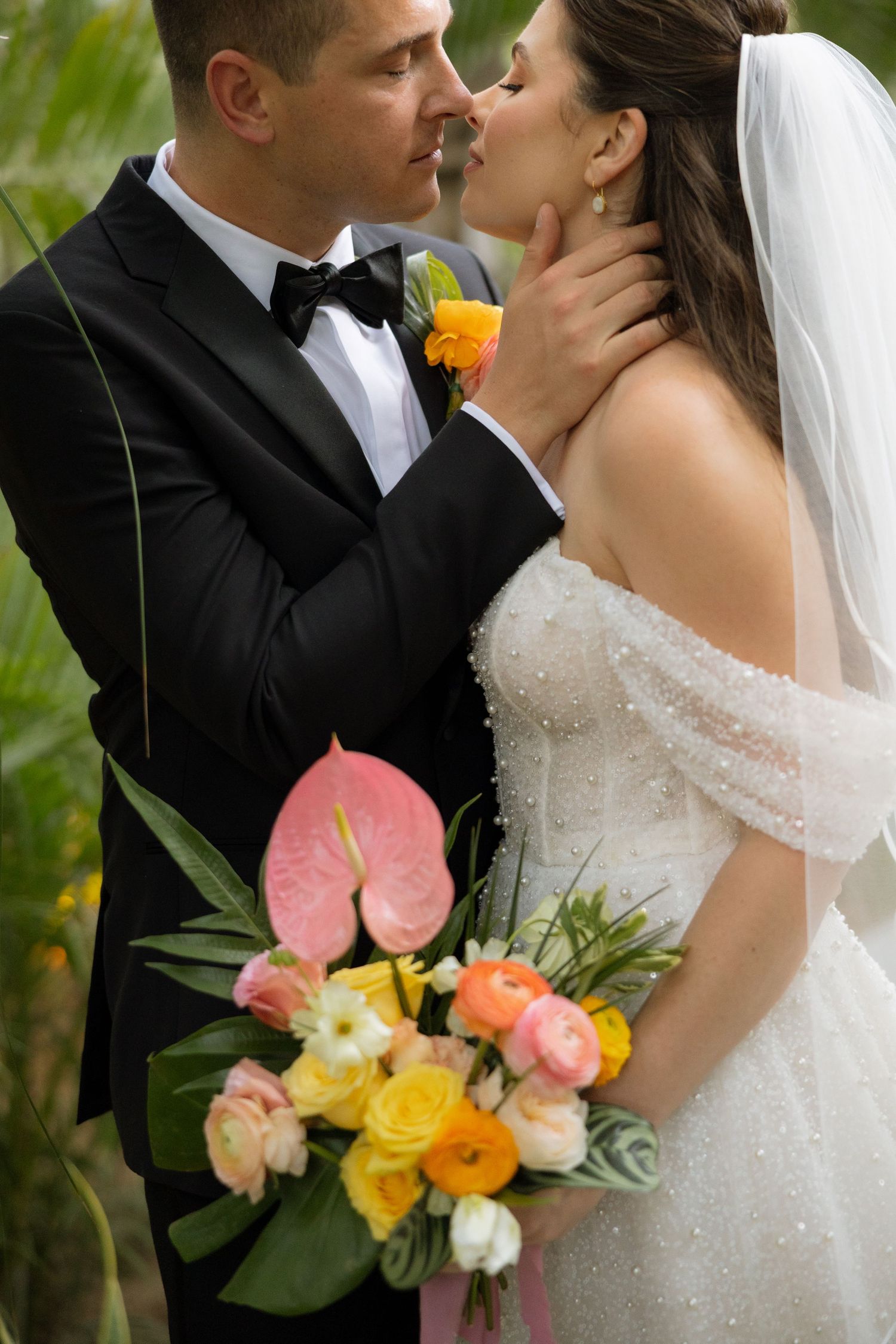 Bride and groom share a romantic kiss, she holds a vibrant bouquet of pink, orange, and yellow flowers.