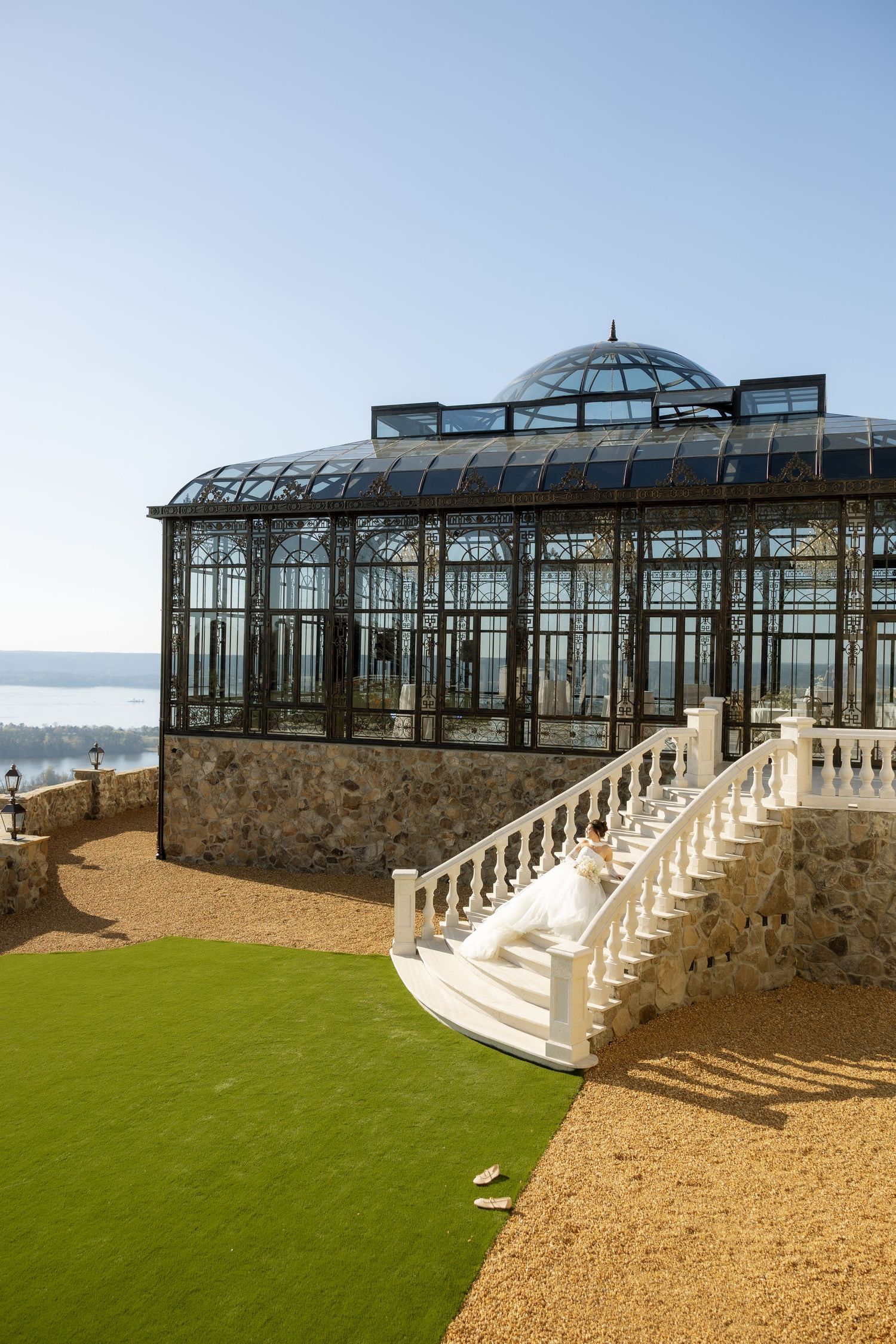 Elegant glass greenhouse conservatory with white balustrade staircase, stone base, and scenic waterfront views under clear blue sky.