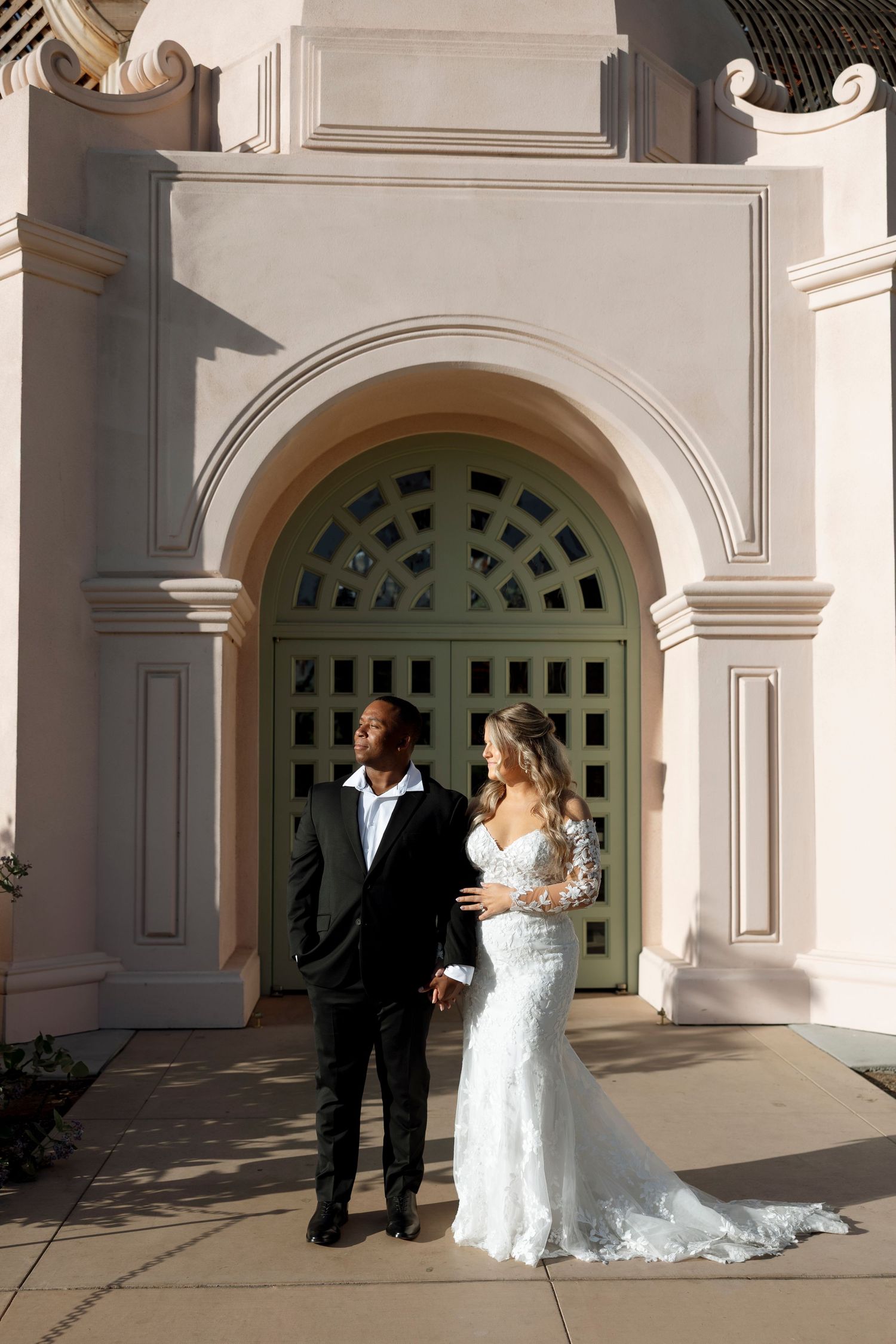 A couple in wedding attire poses in front of an elegant arched doorway with green glass panels.