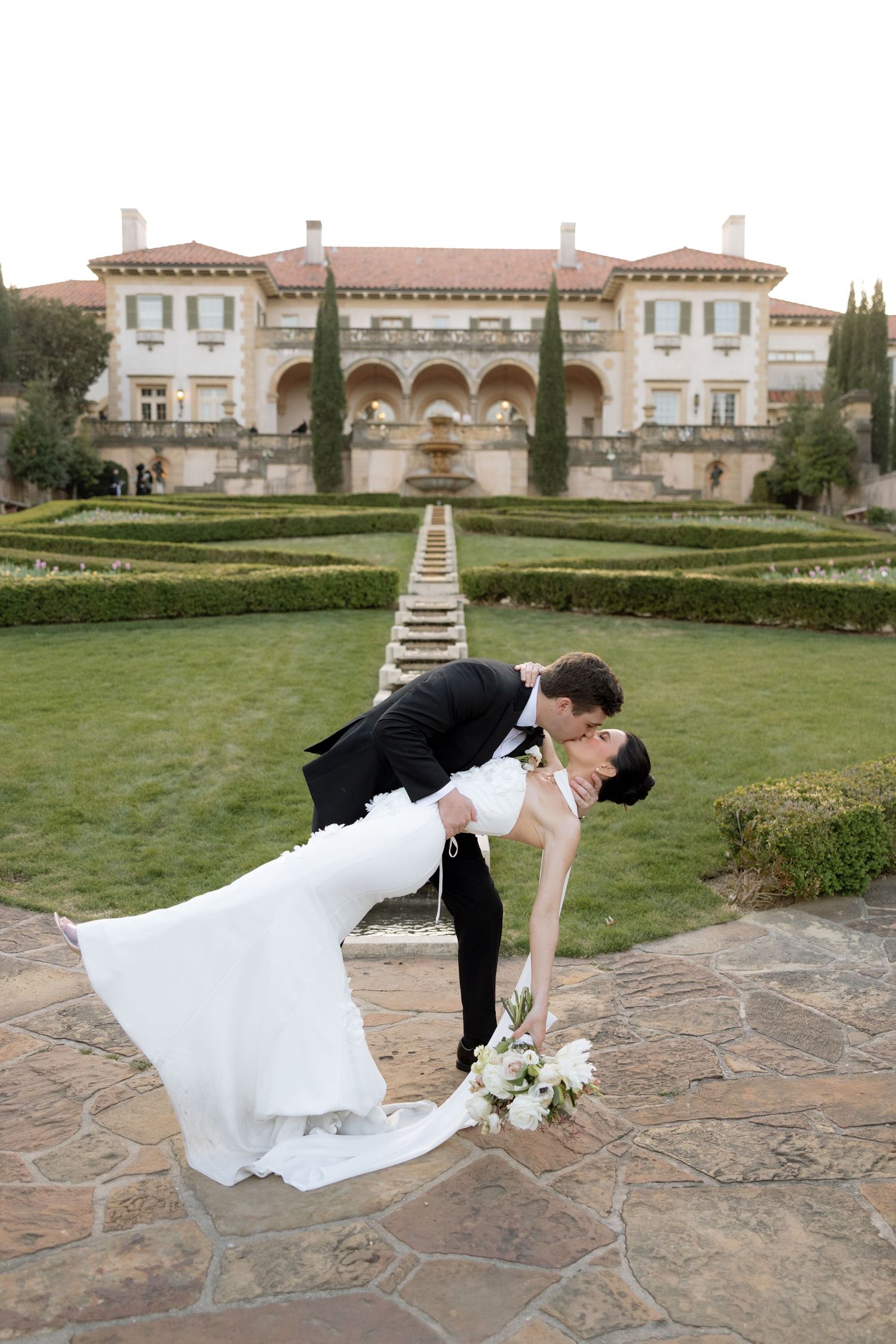 Groom dips bride in white gown before grand Italian-style mansion with manicured gardens.