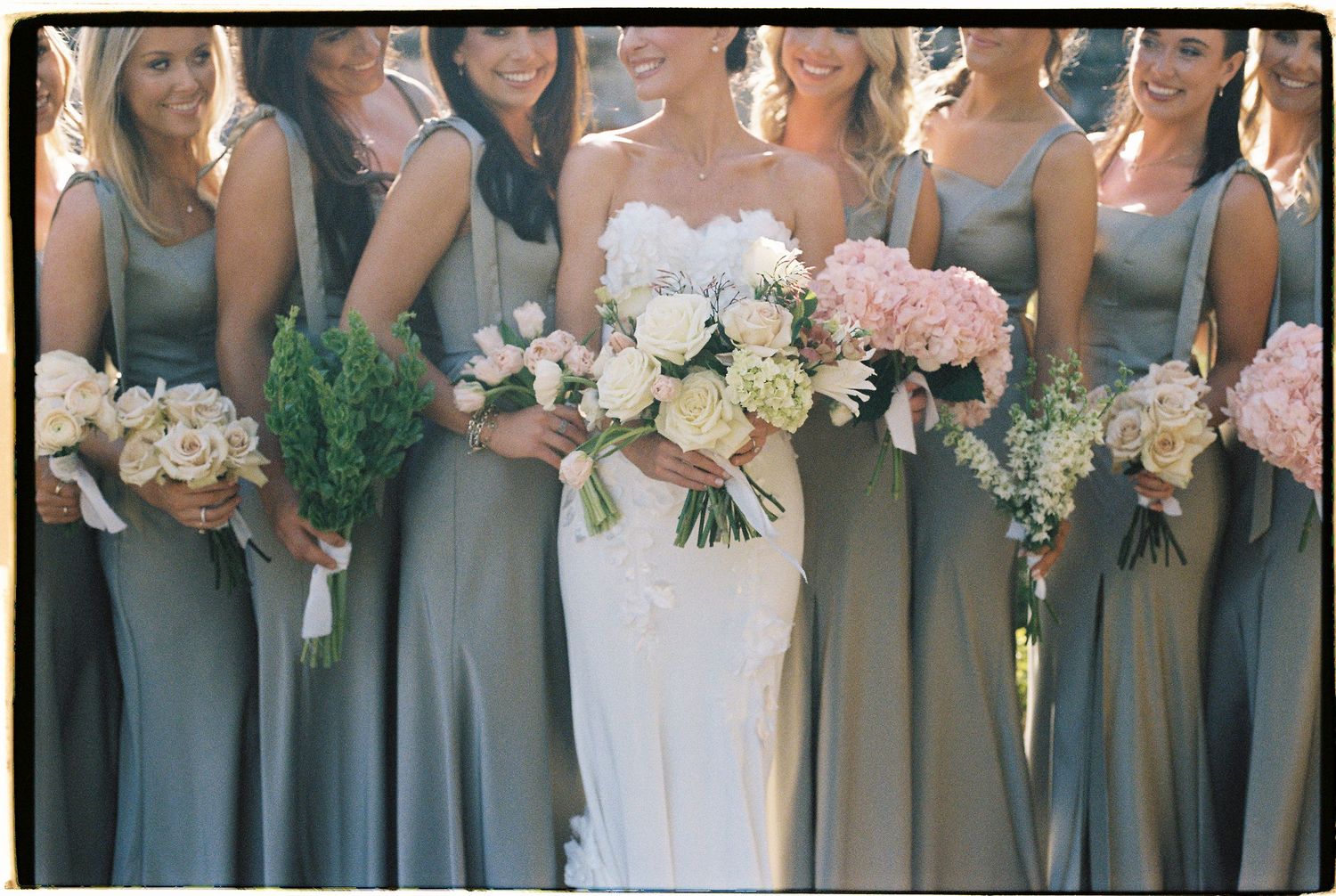 Bride in white gown with bridesmaids in sage green dresses holding pink and white floral bouquets at wedding.