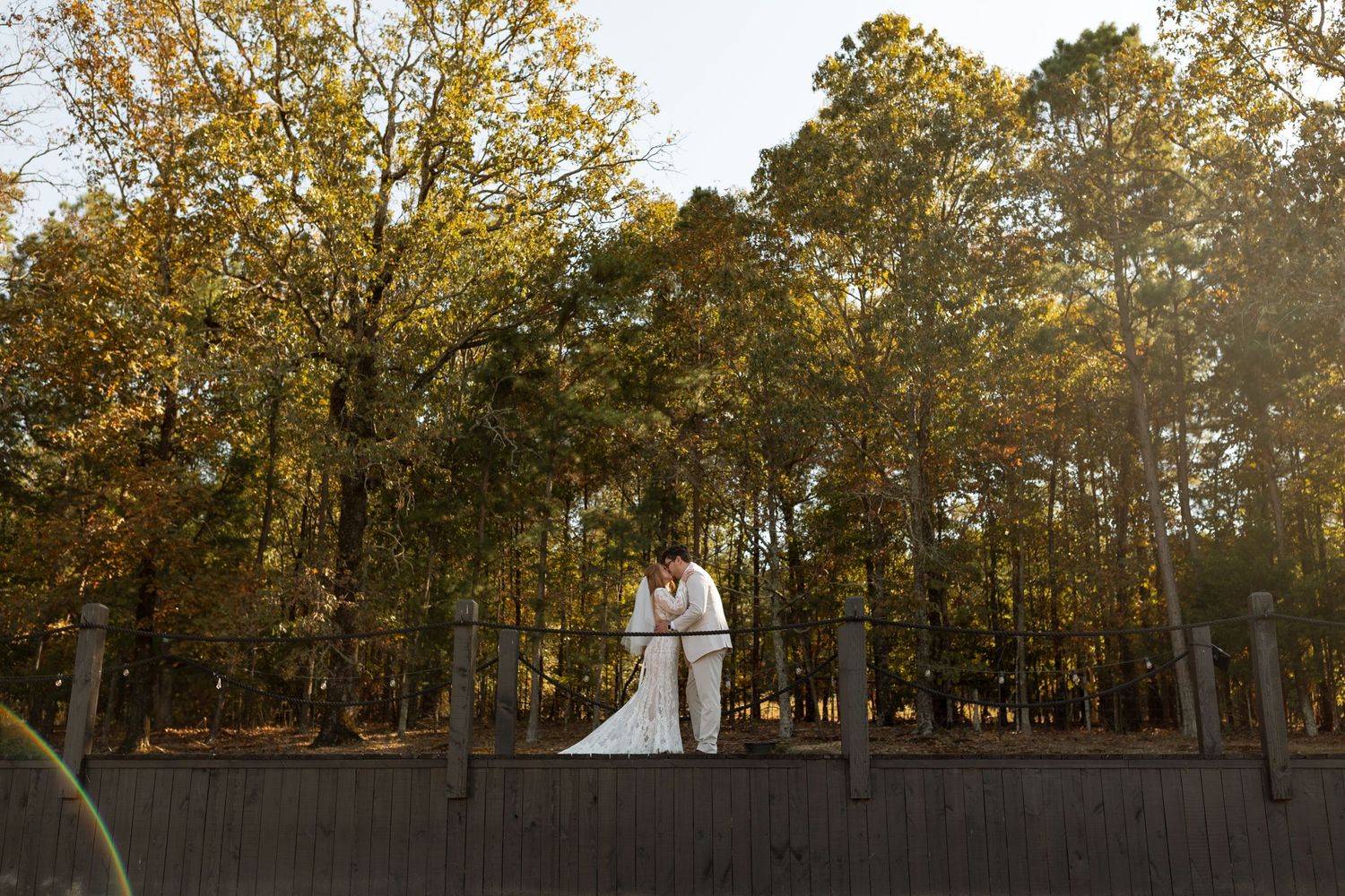 Wedding couple standing on wooden deck surrounded by autumn trees at sunset.