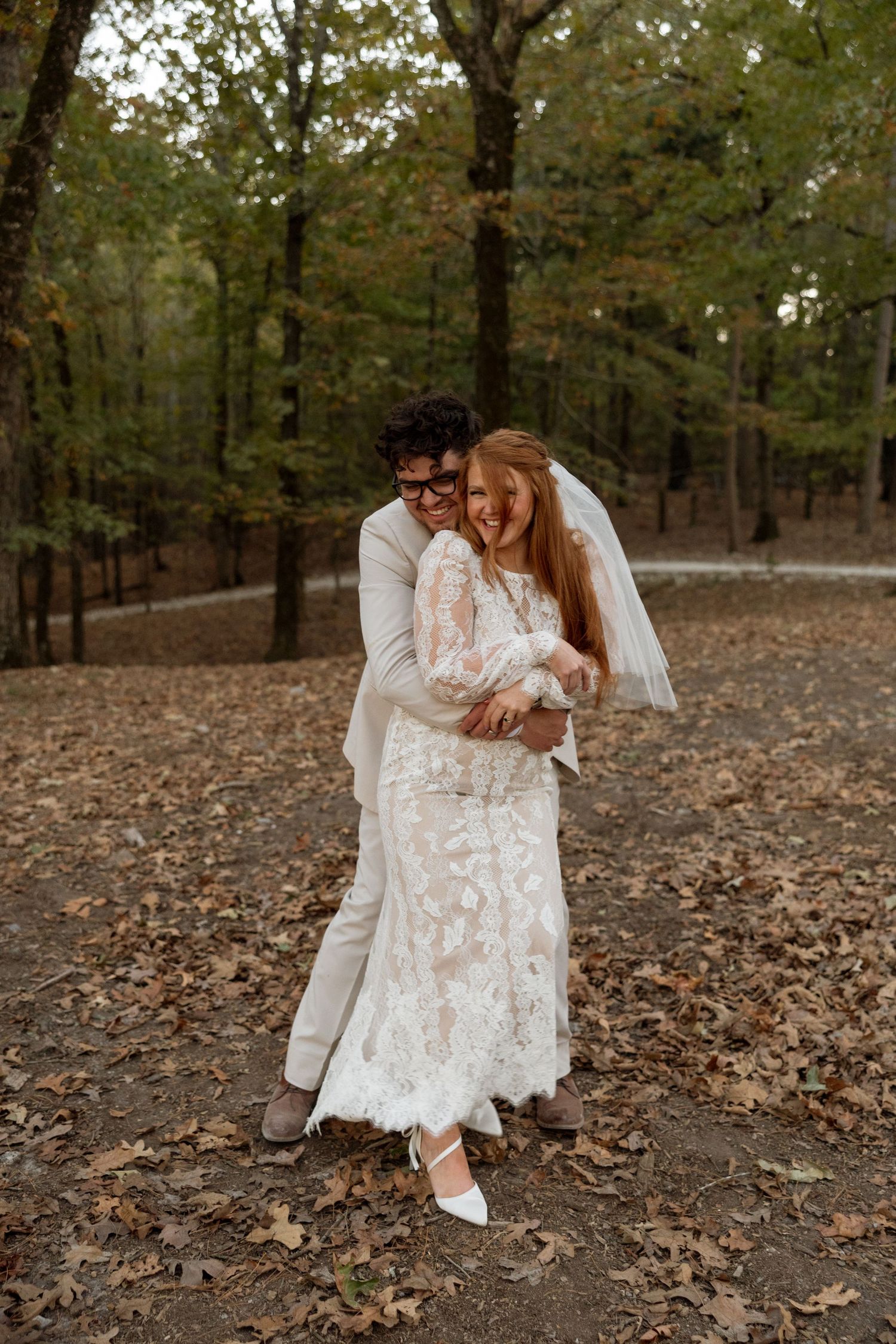 Joyful newlyweds share a laugh while posing in a wooded area covered with fallen autumn leaves.
