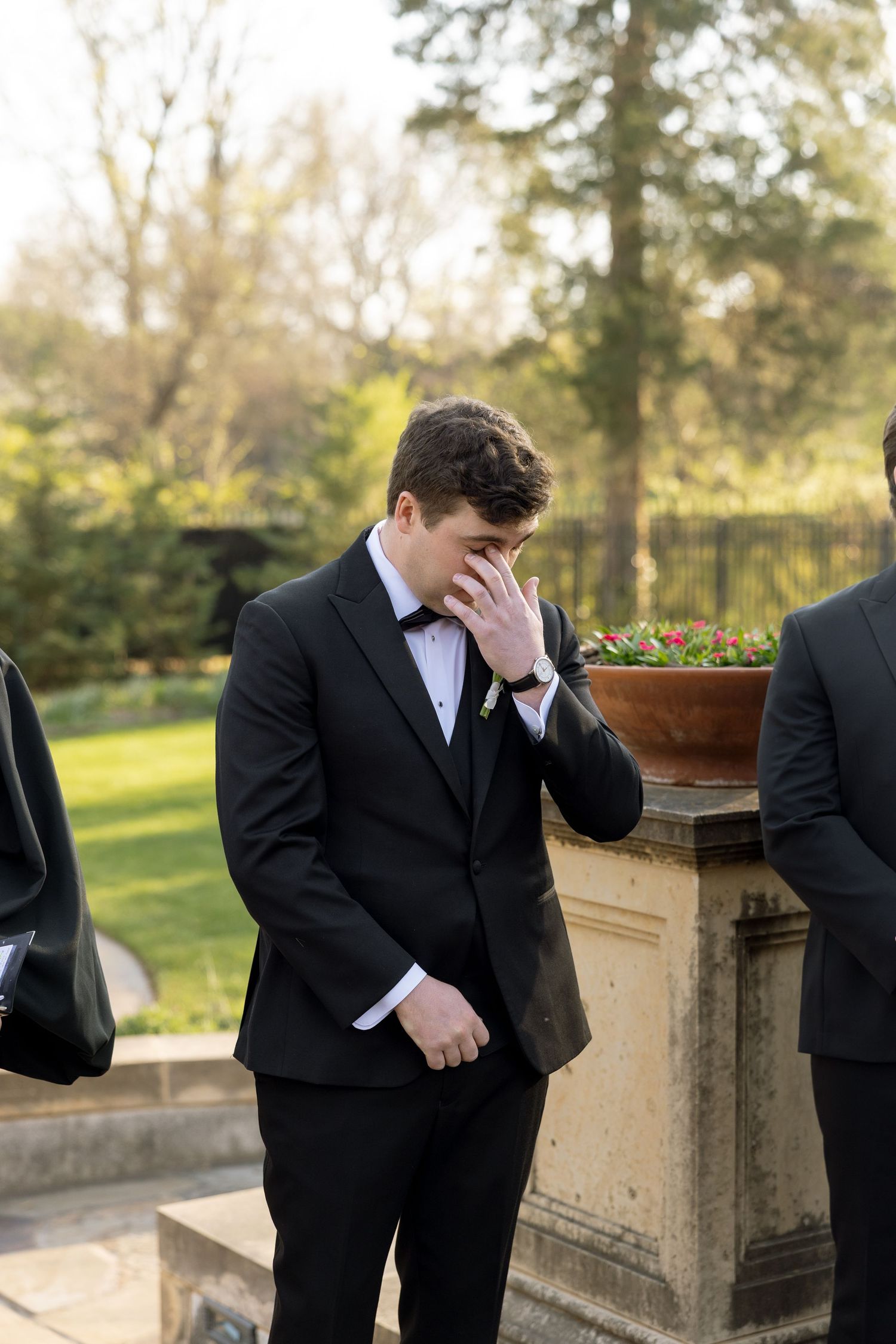 Emotional groom wiping tears in black tuxedo with boutonniere during outdoor wedding ceremony.