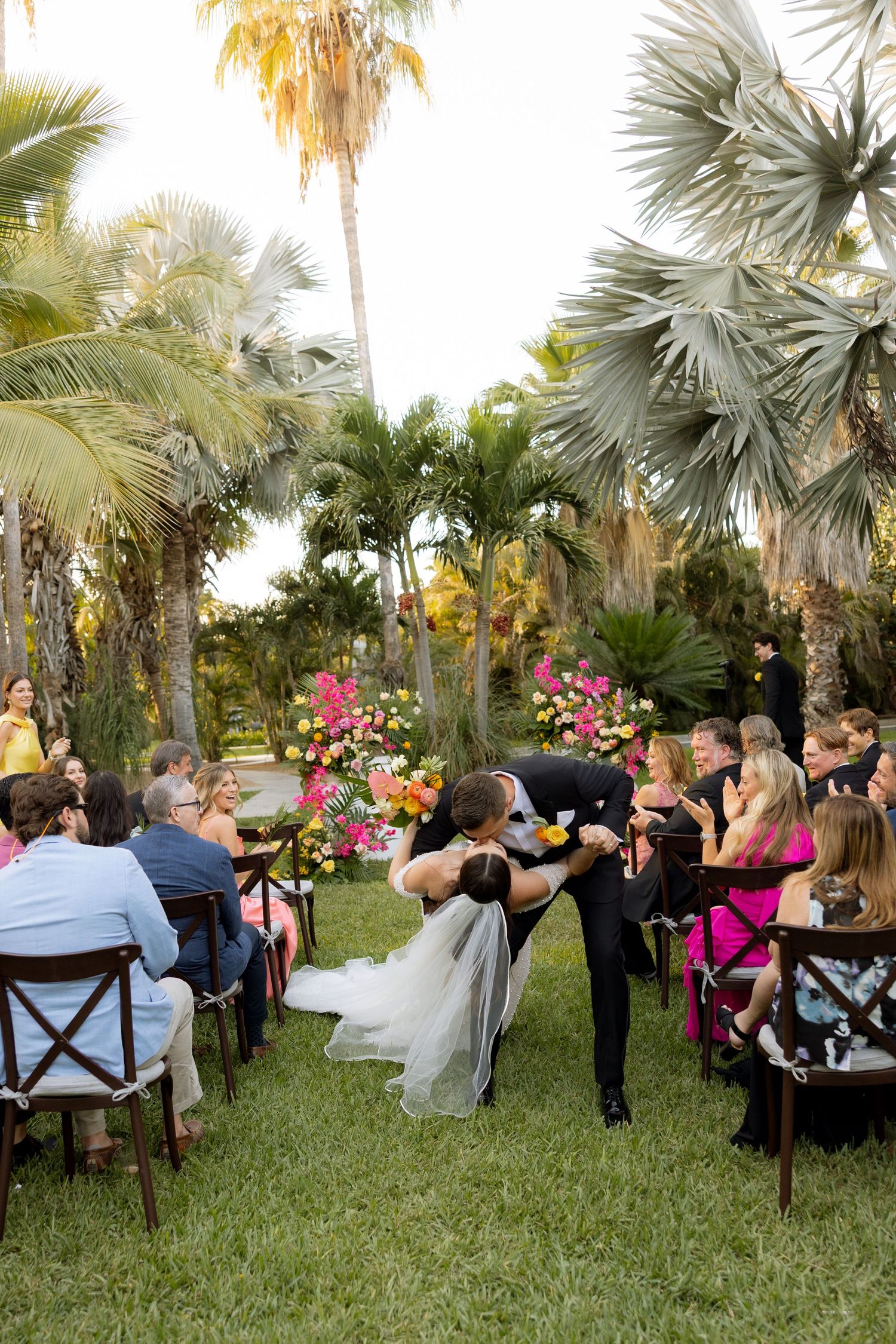 Groom dips bride at outdoor tropical wedding ceremony while guests applaud in background.