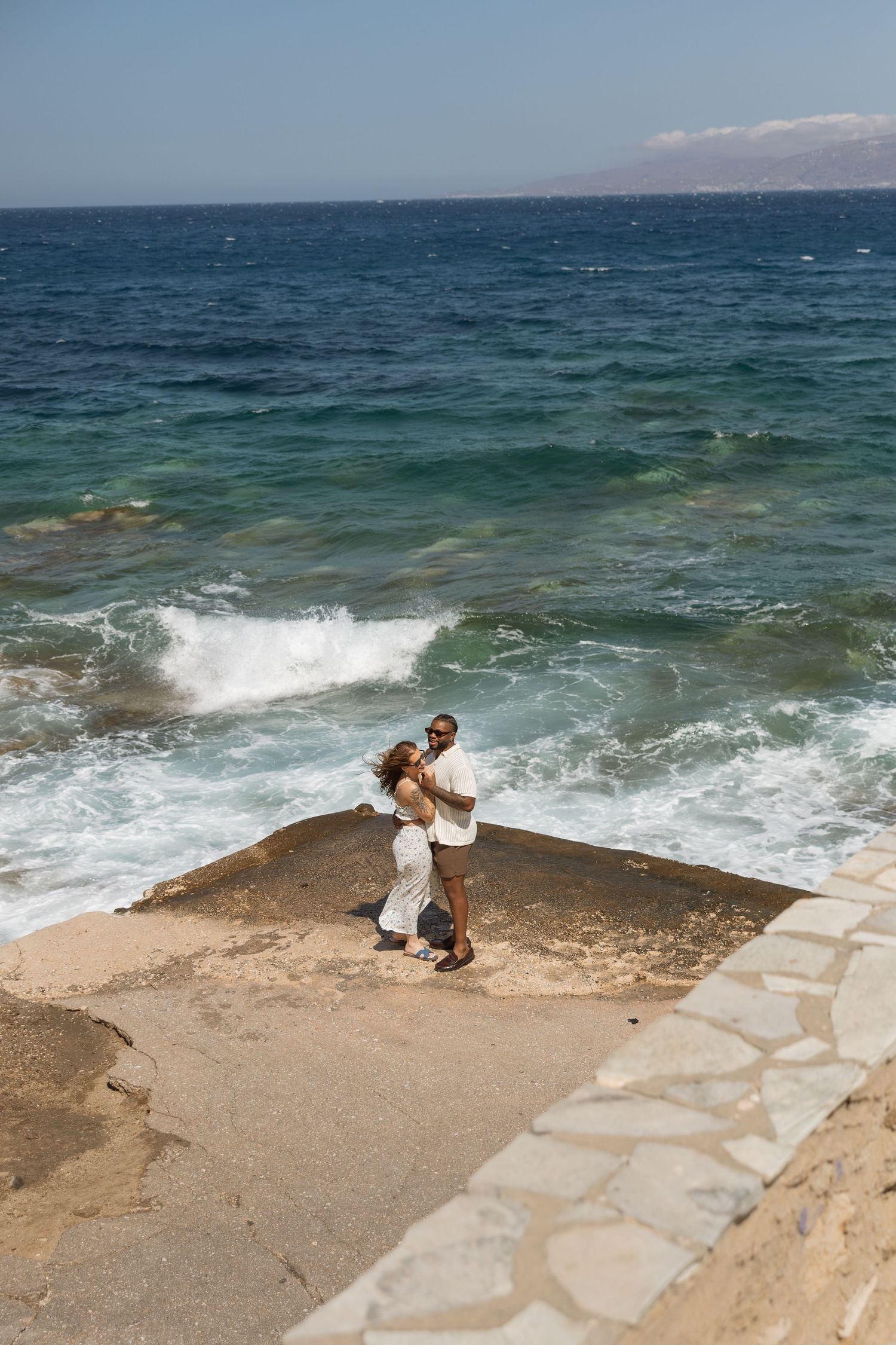 A couple embraces on a stone platform overlooking the Mediterranean Sea with crashing waves in the background.