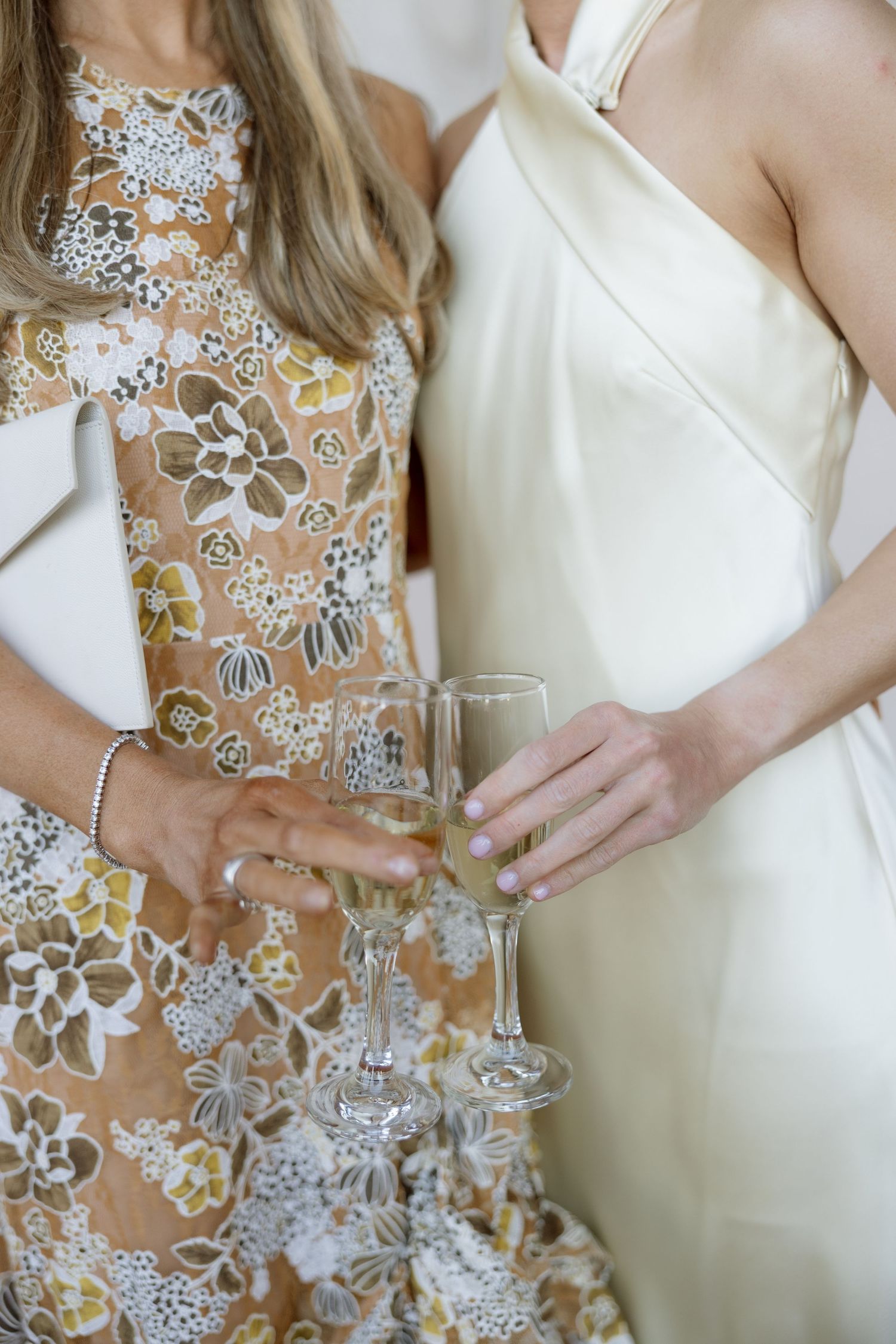 Two women in elegant dresses clinking champagne glasses at a wedding celebration.