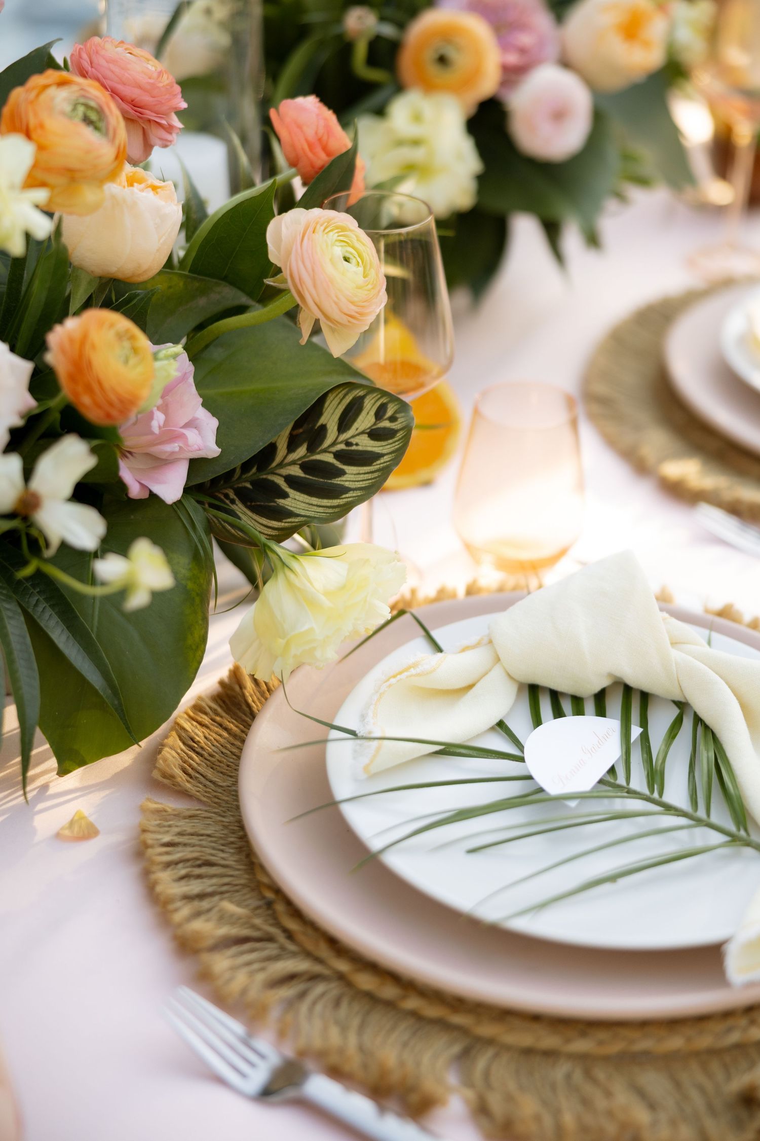 Elegant tropical wedding table setting with ranunculus flowers, palm leaves, and gold charger plates.