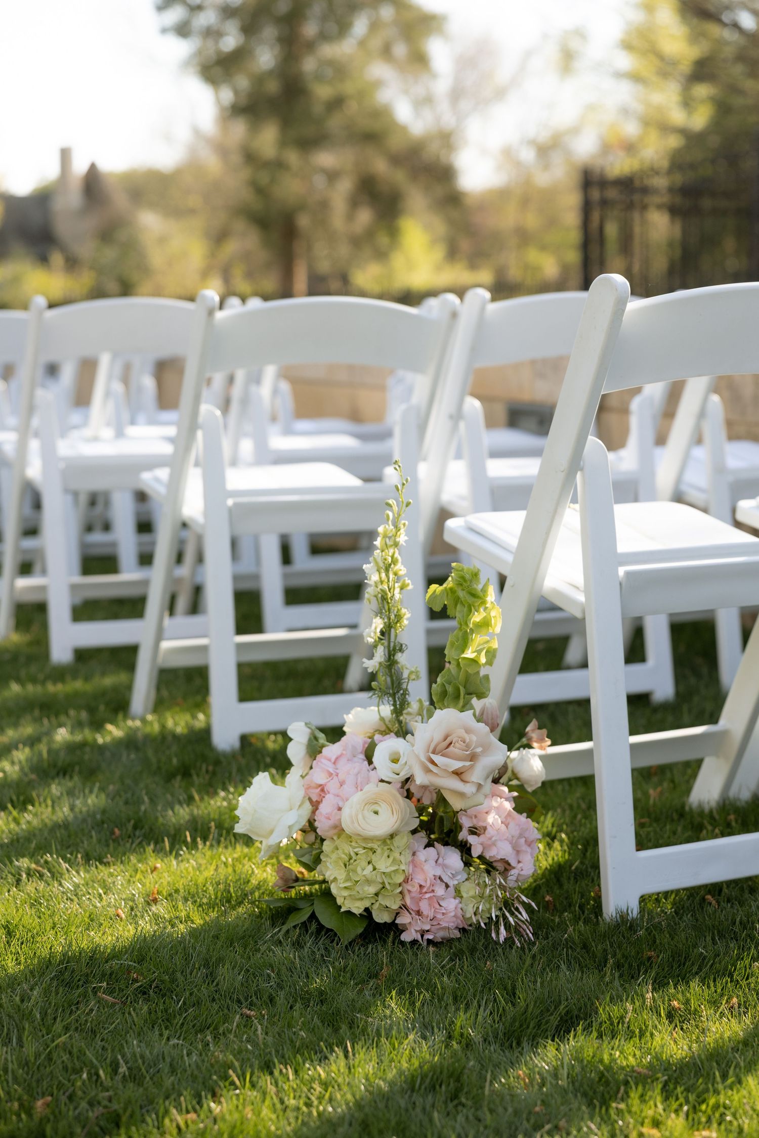 Pink and white floral bouquet with hydrangeas and roses adorns aisle of white folding chairs at outdoor wedding.
