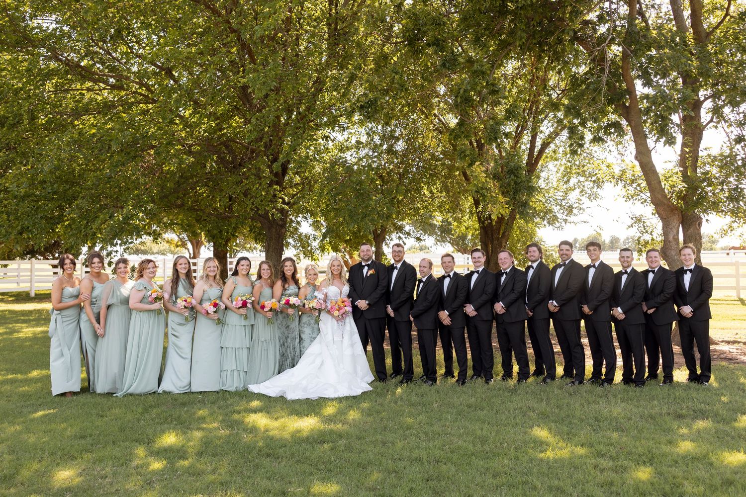 Wedding party poses outdoors in a line formation against a backdrop of green trees.