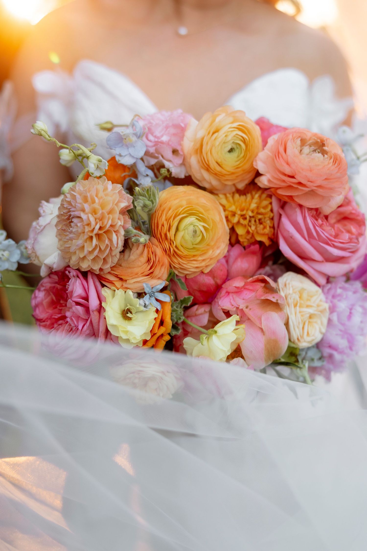 Delicate floral arrangement featuring pink, peach and yellow ranunculus and roses against soft fabric.