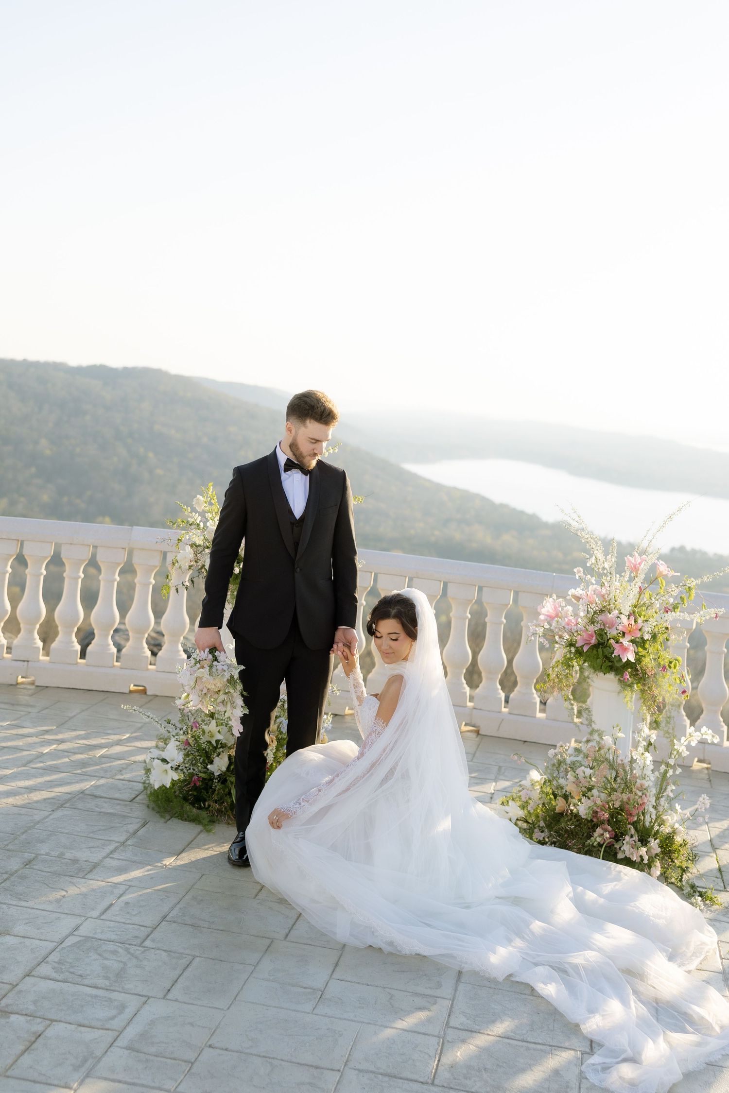 Bride in white gown and groom in black tuxedo pose on scenic terrace with floral arrangements and mountain lake view.