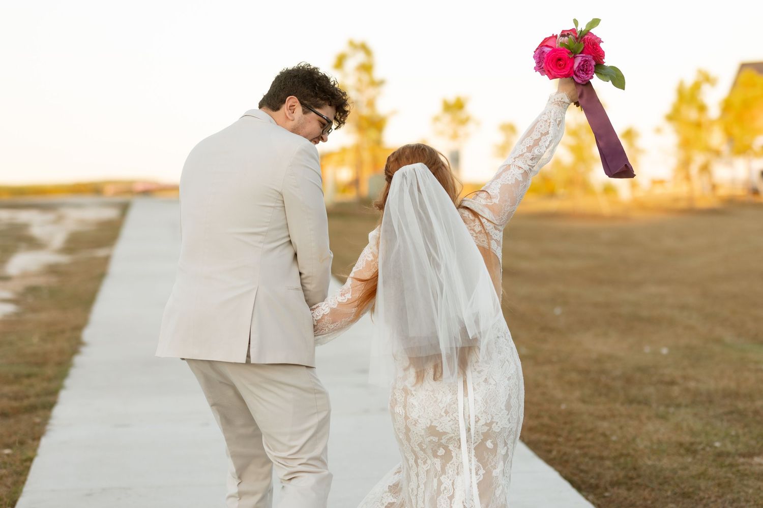 Couple walking on path at sunset with raised bouquet in celebratory pose.
